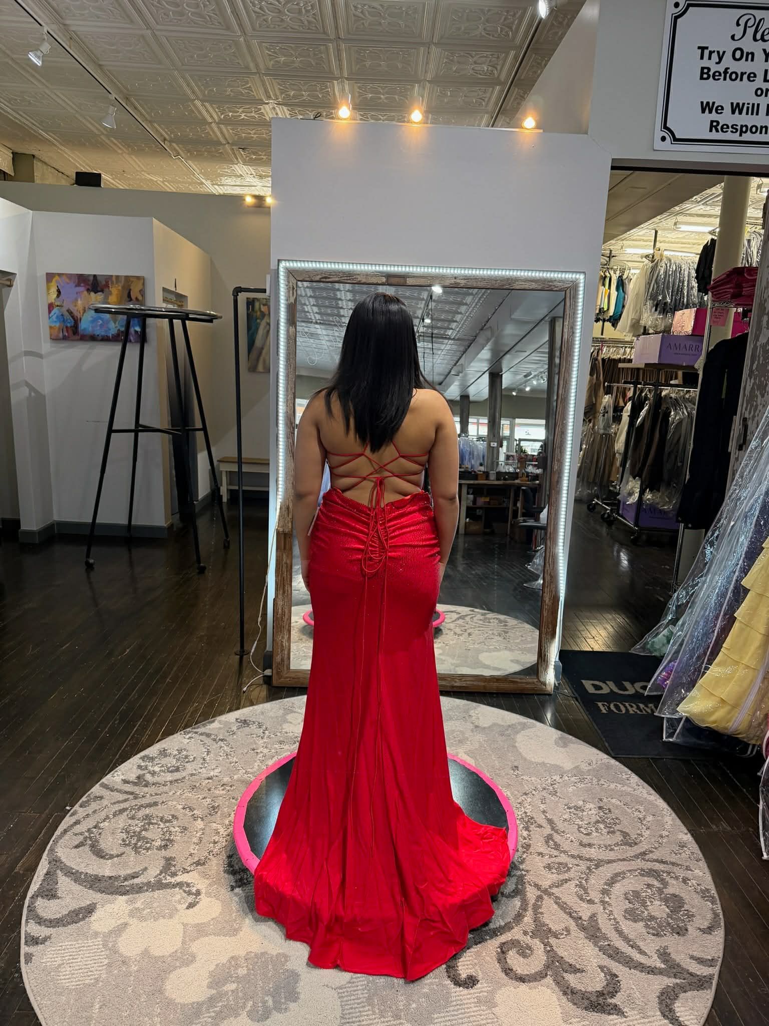 A person stands on a small circular platform in a dressing room, wearing a floor-length red gown with an open back.