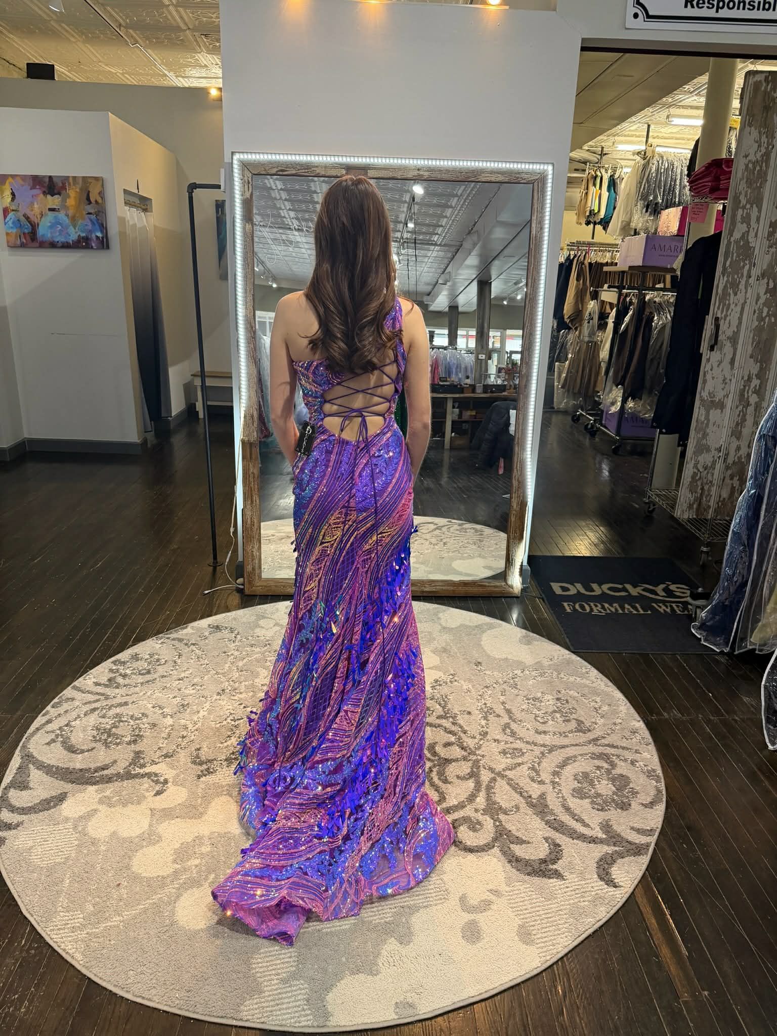 A person stands in a dress shop wearing a sparkling purple, pink, and blue gown with a lace-up back, viewed from behind.