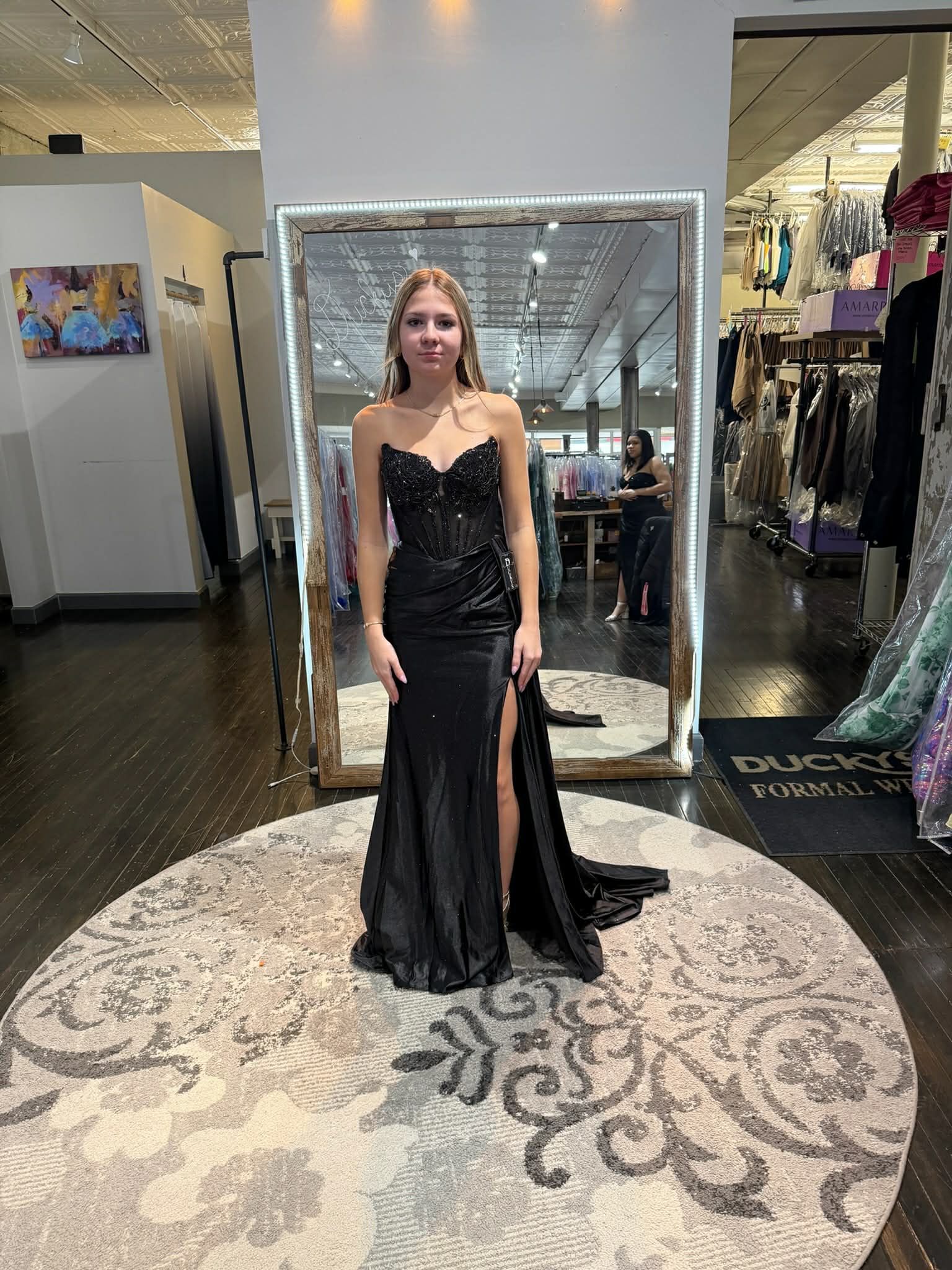 A person stands in a dress shop wearing a black, strapless, floor-length gown with a side slit, reflected in a large mirror.