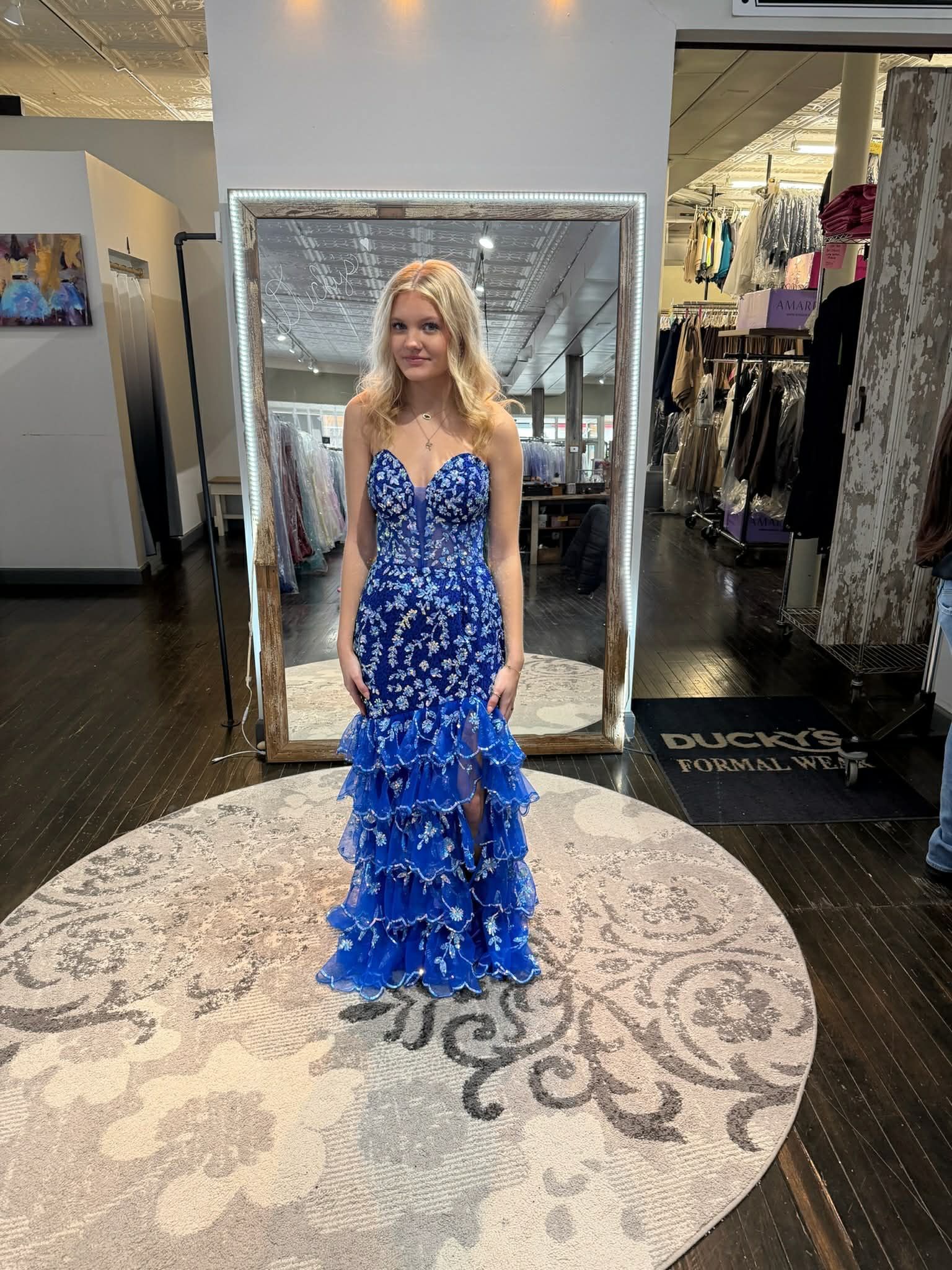 A person wearing a floor-length blue, floral-patterned dress with a tiered skirt stands on a circular rug in a store.