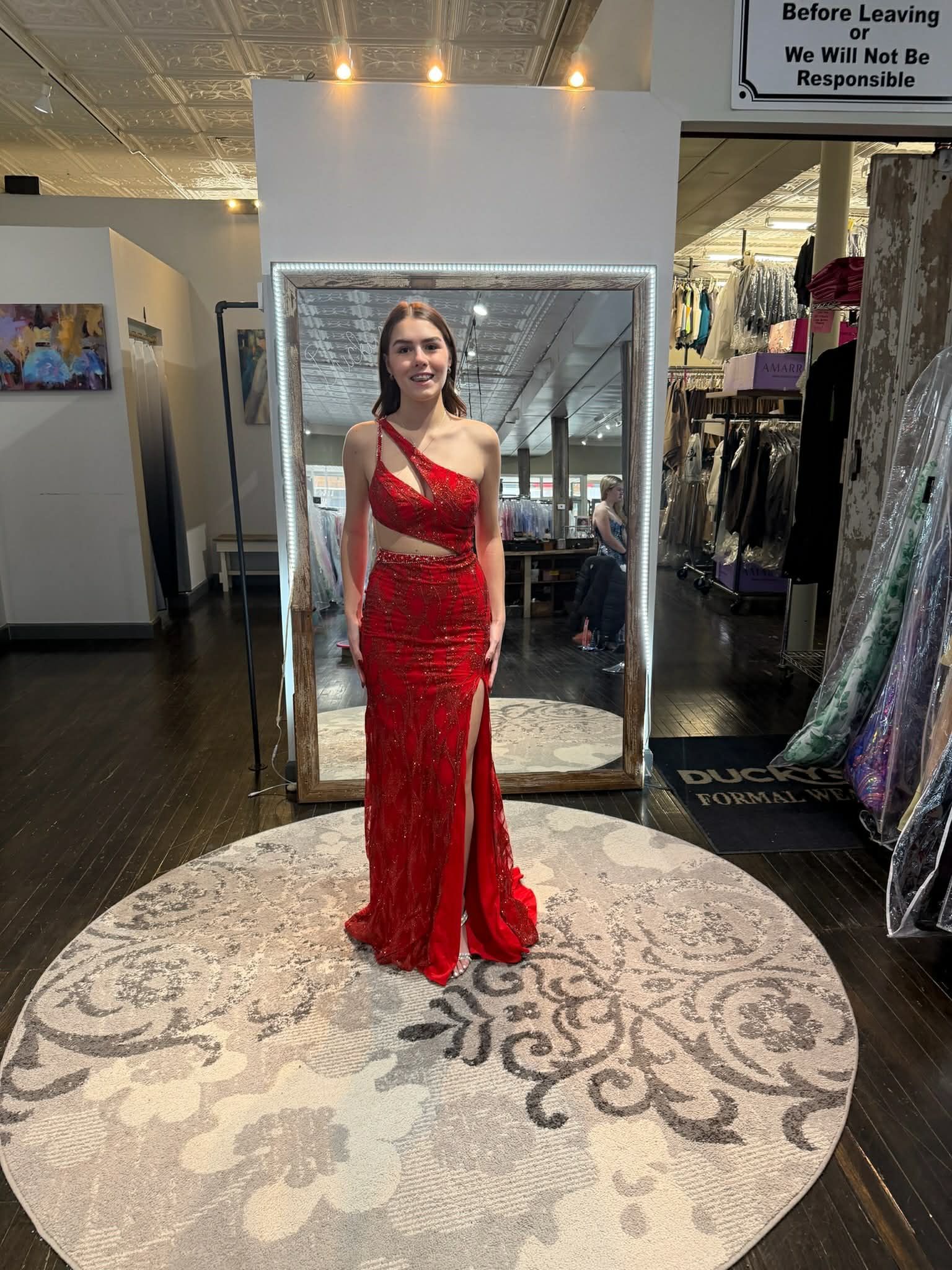 A person stands in a dress shop, modeling a floor-length, red sequined one-shoulder gown with a cutout and side slit.