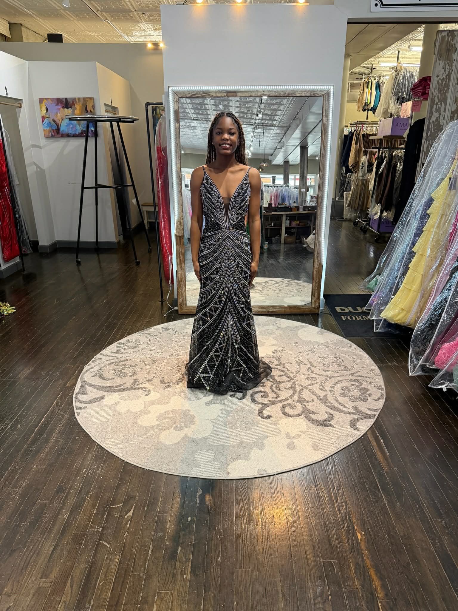 A person stands centered on a circular rug in a formal wear boutique, wearing a floor-length, black and silver sequined gown.