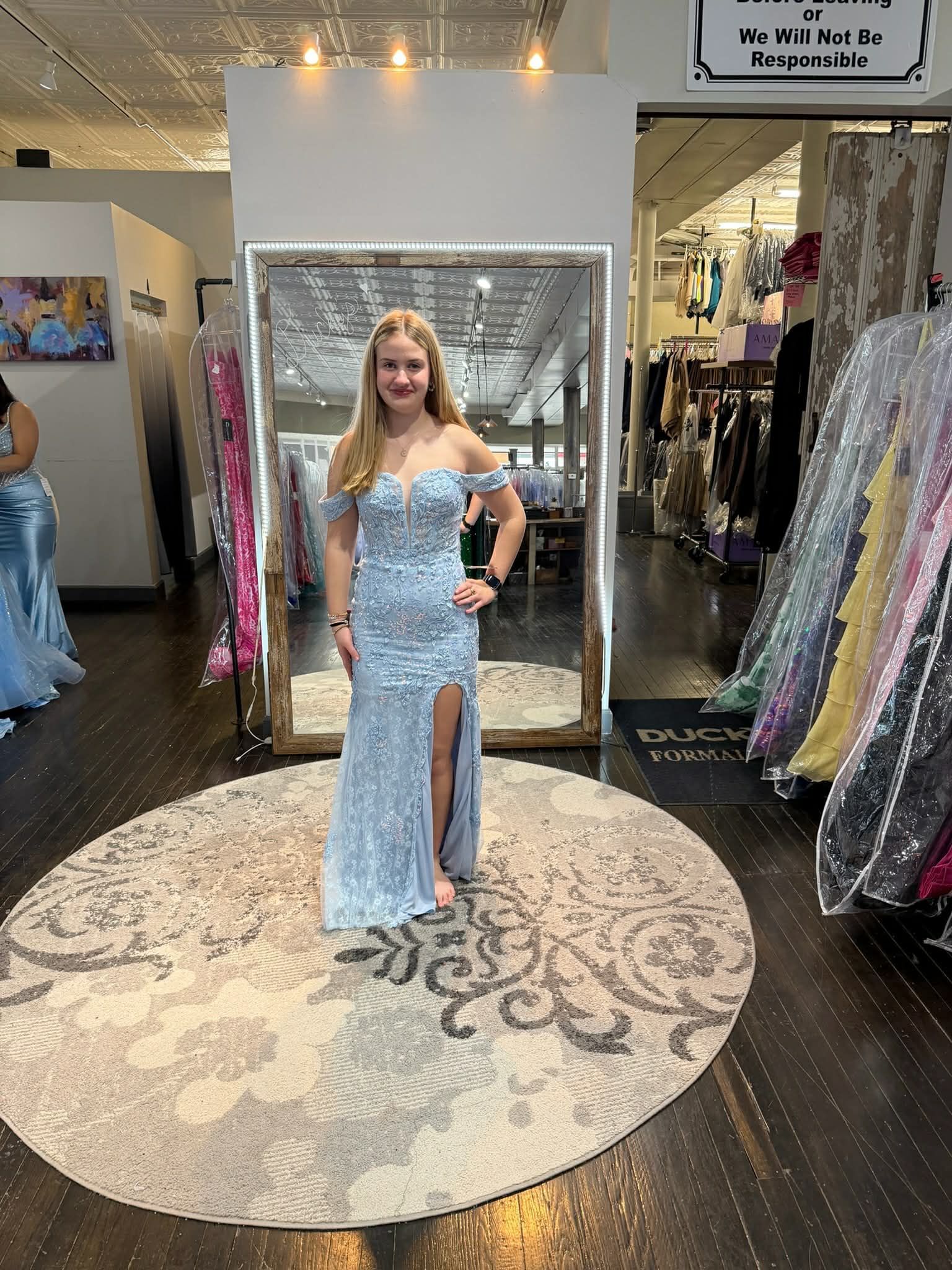 A person wearing a light blue, floor-length floral lace gown with an off-the-shoulder neckline stands in a dress shop.