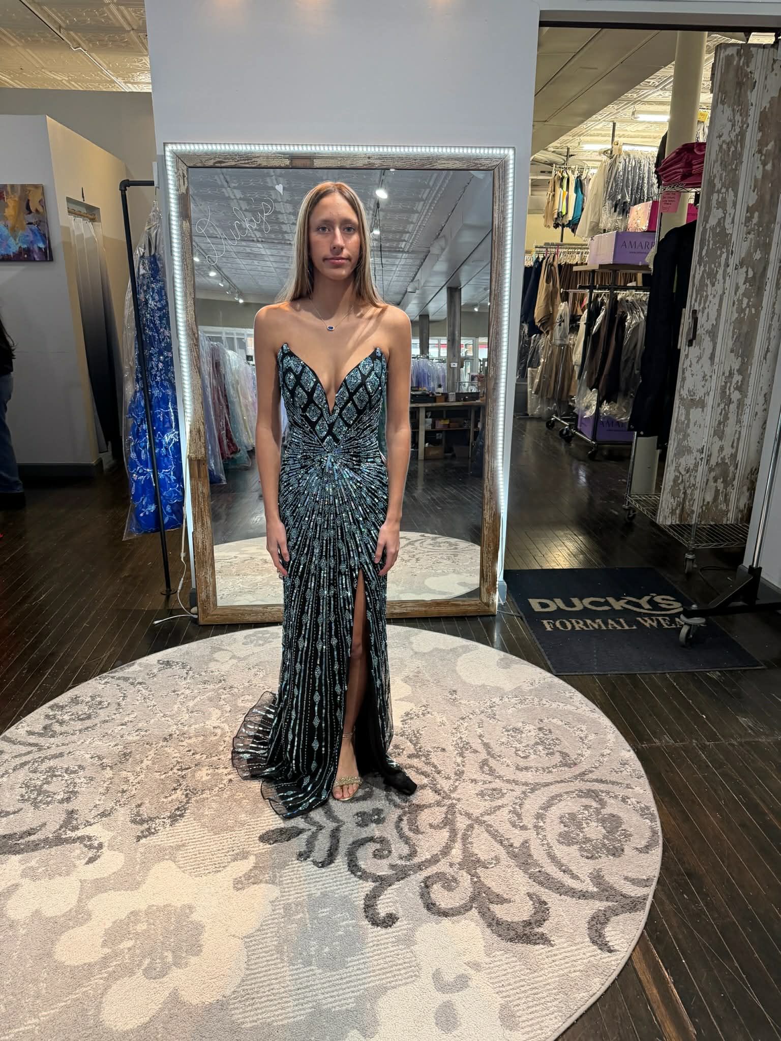 A person stands on a circular rug in a boutique, wearing a floor-length, dark, sequined evening gown with a thigh-high slit.