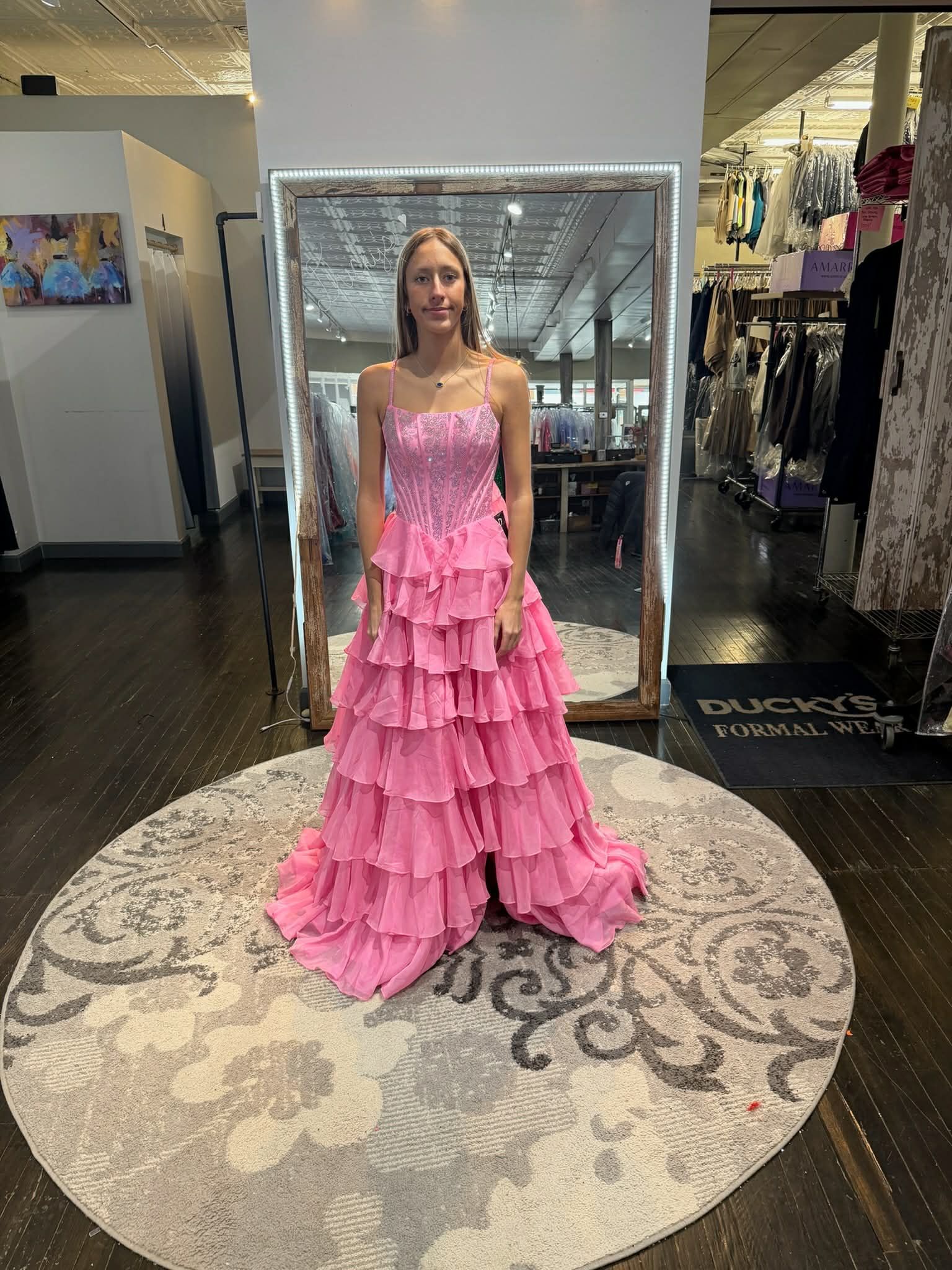 A person stands in a clothing store wearing a vibrant pink, floor-length tiered ruffle dress with a beaded bodice.