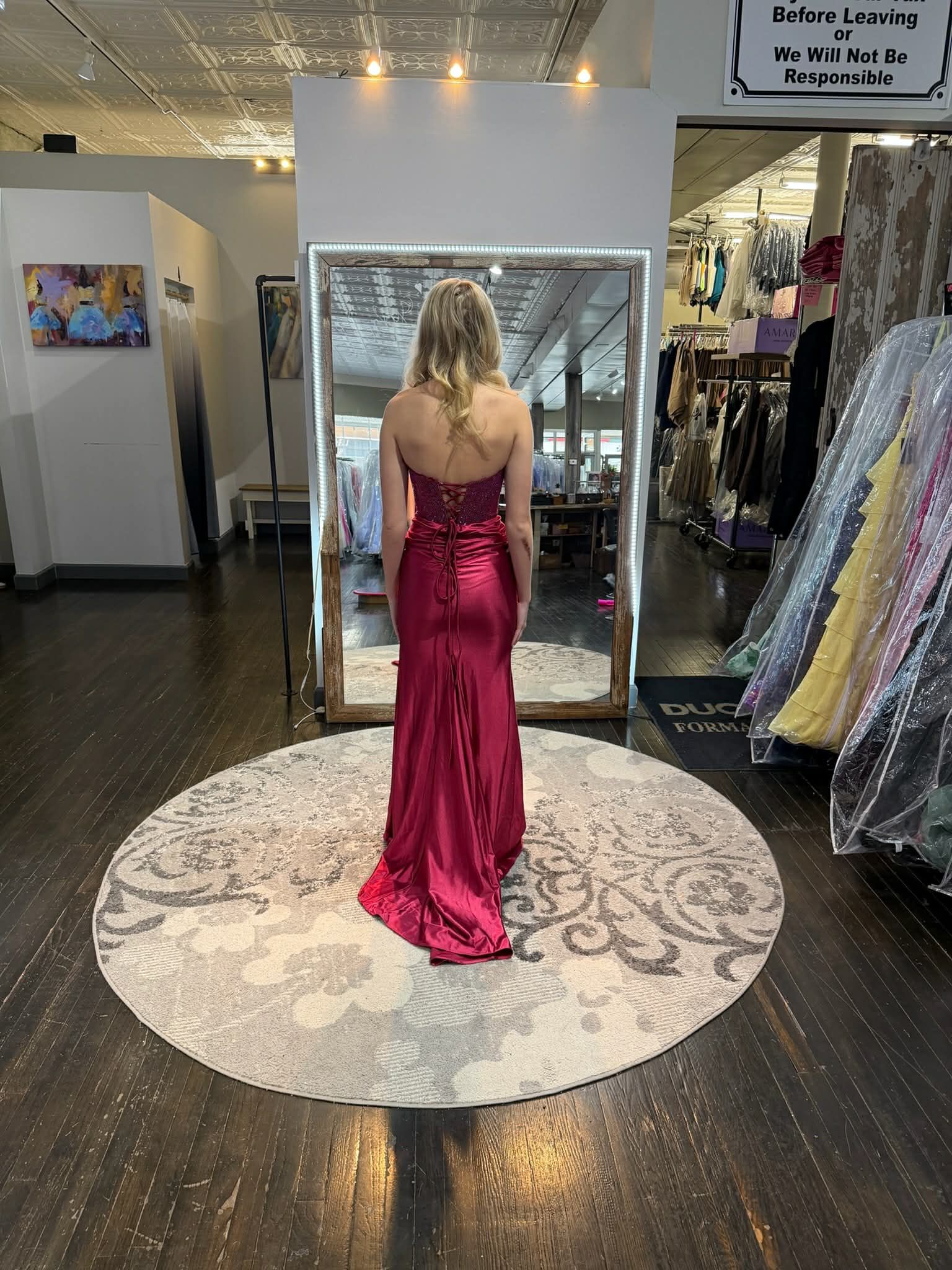 A person wearing a floor-length, fuchsia satin gown stands in front of a floor mirror inside a clothing store.