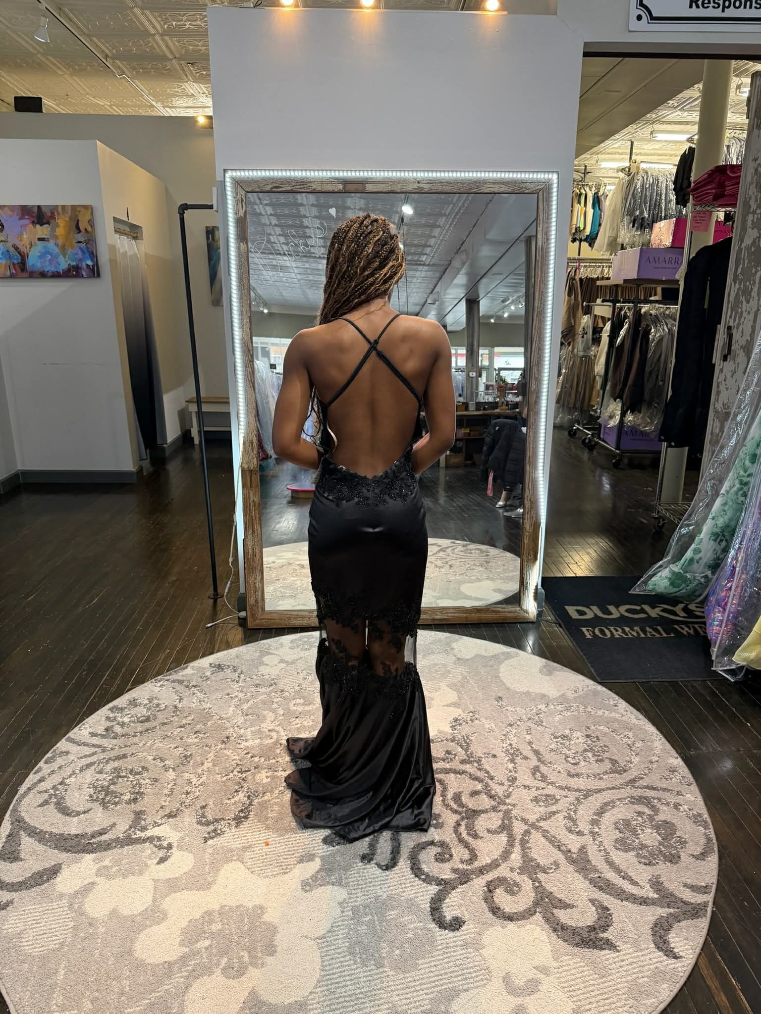 A person standing on a round rug in a clothing store, facing a mirror while wearing a black, backless, floor-length dress.