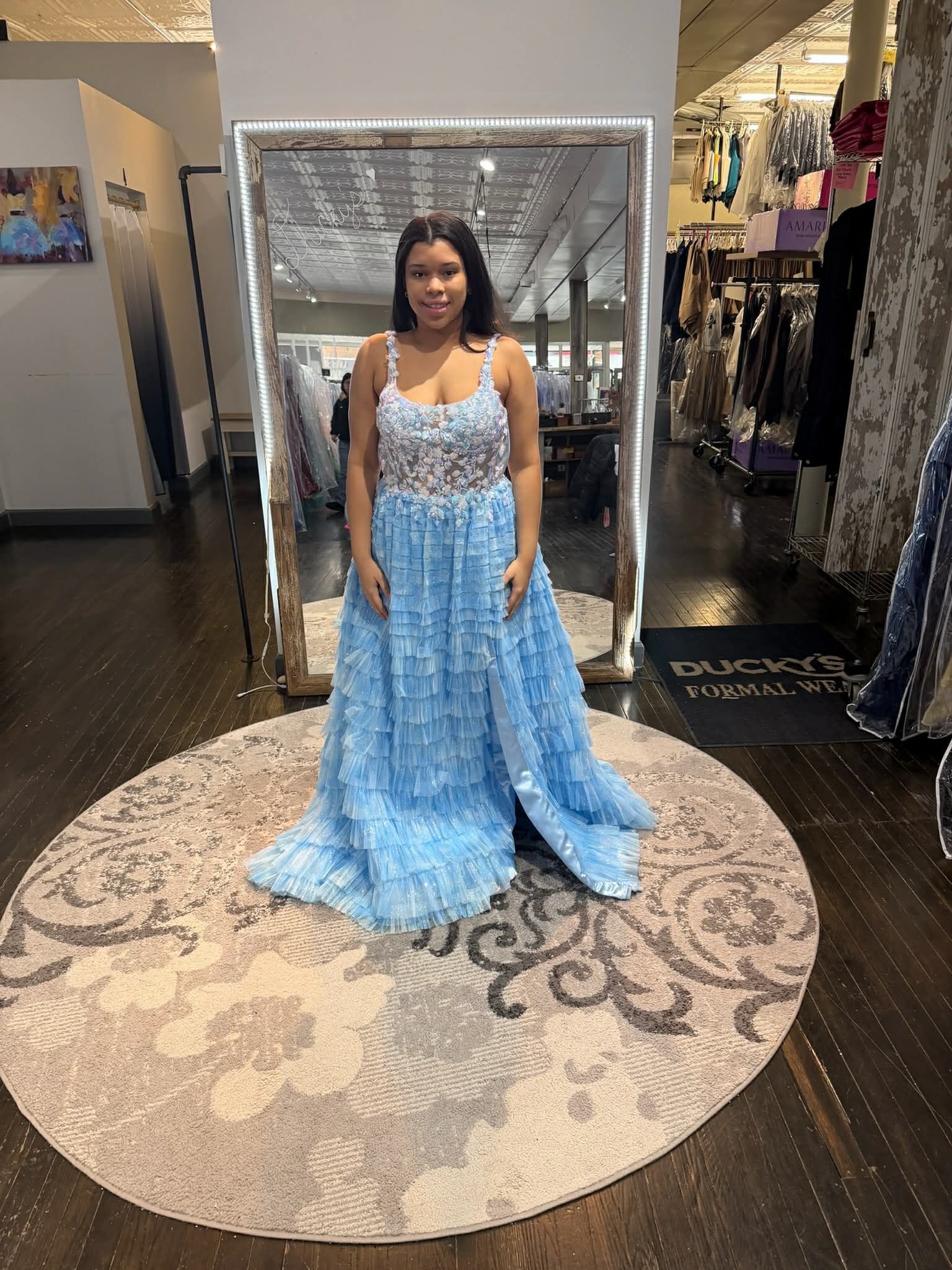 A person wearing a light blue formal gown with a sequined bodice and floral skirt stands on a rug in a dress shop.