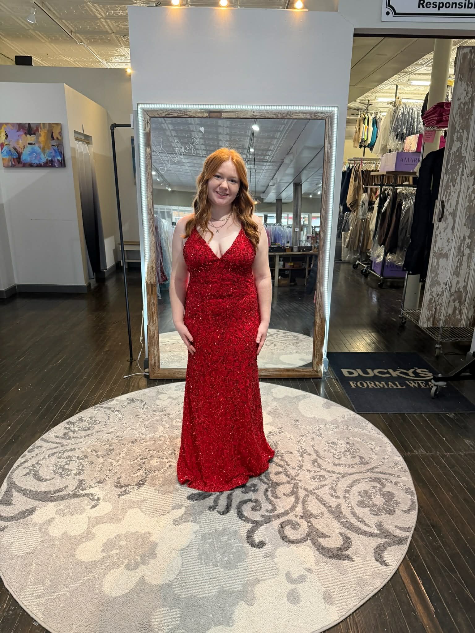 A smiling person stands in a dress shop wearing a floor-length, shimmering red gown in front of a large mirror.