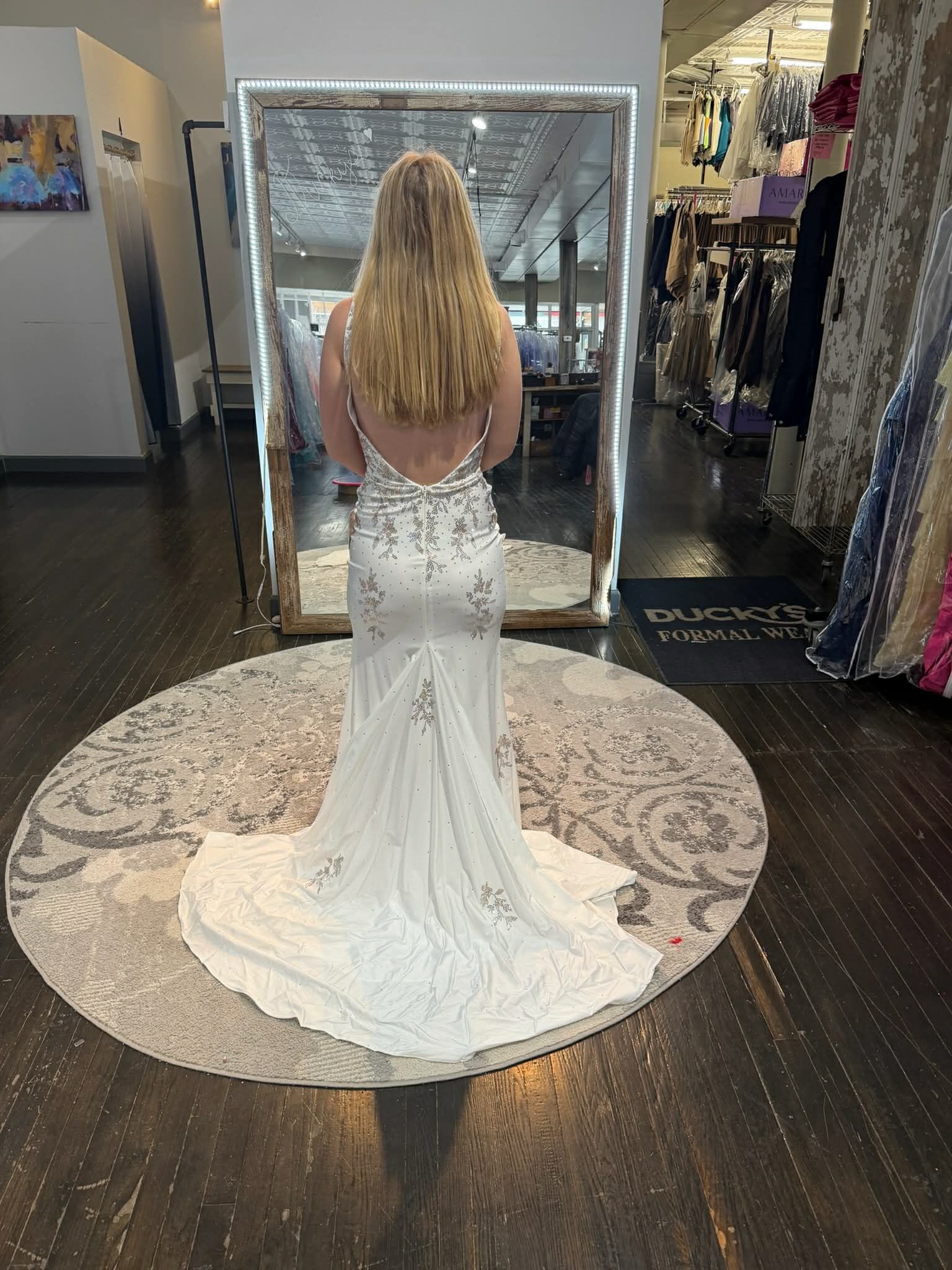A person with long blonde hair viewed from behind, wearing a form-fitting white gown with a train in a clothing store.