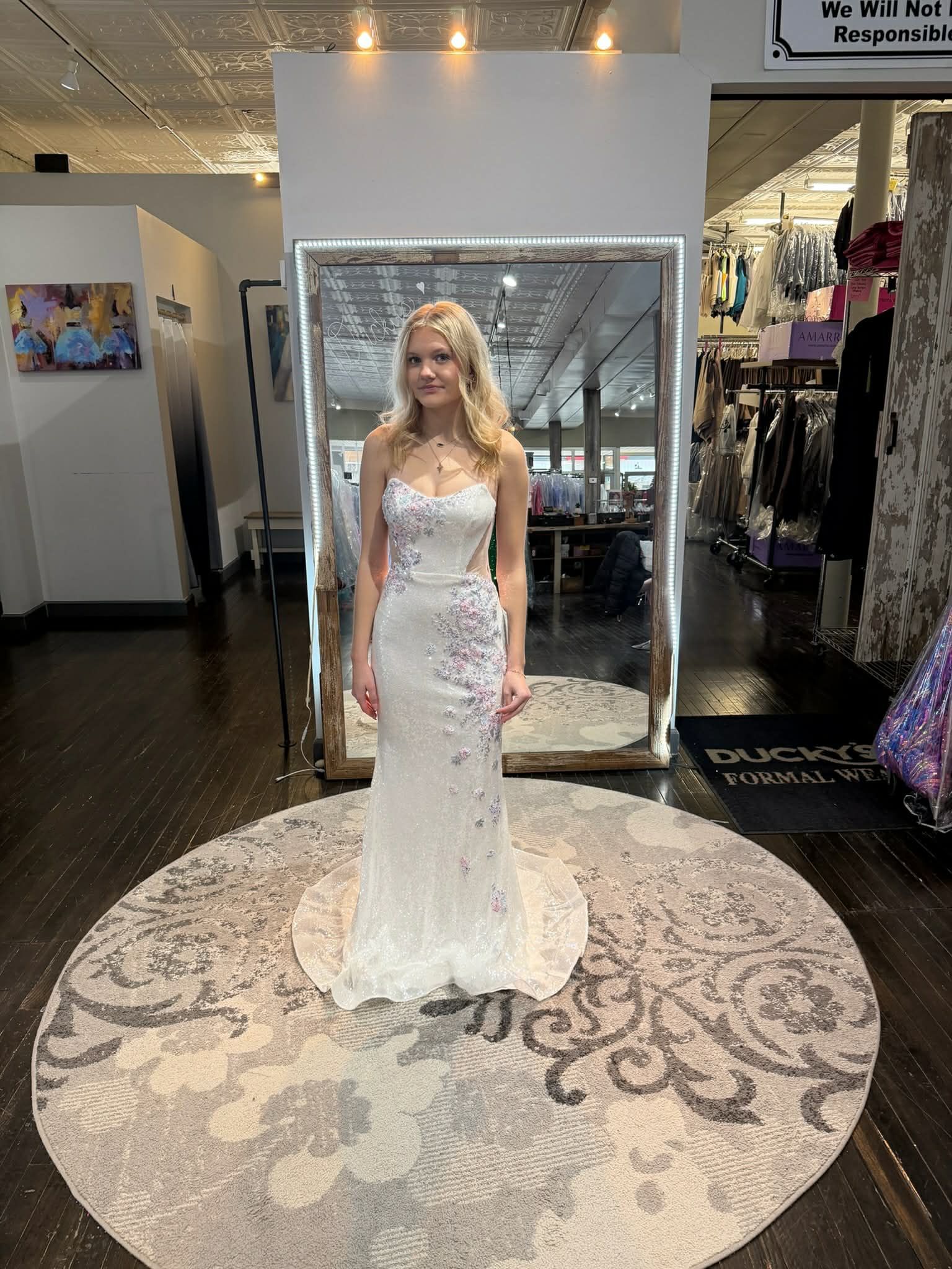 A person wearing a floor-length white, beaded gown stands in a boutique on a patterned circular rug in front of a mirror.