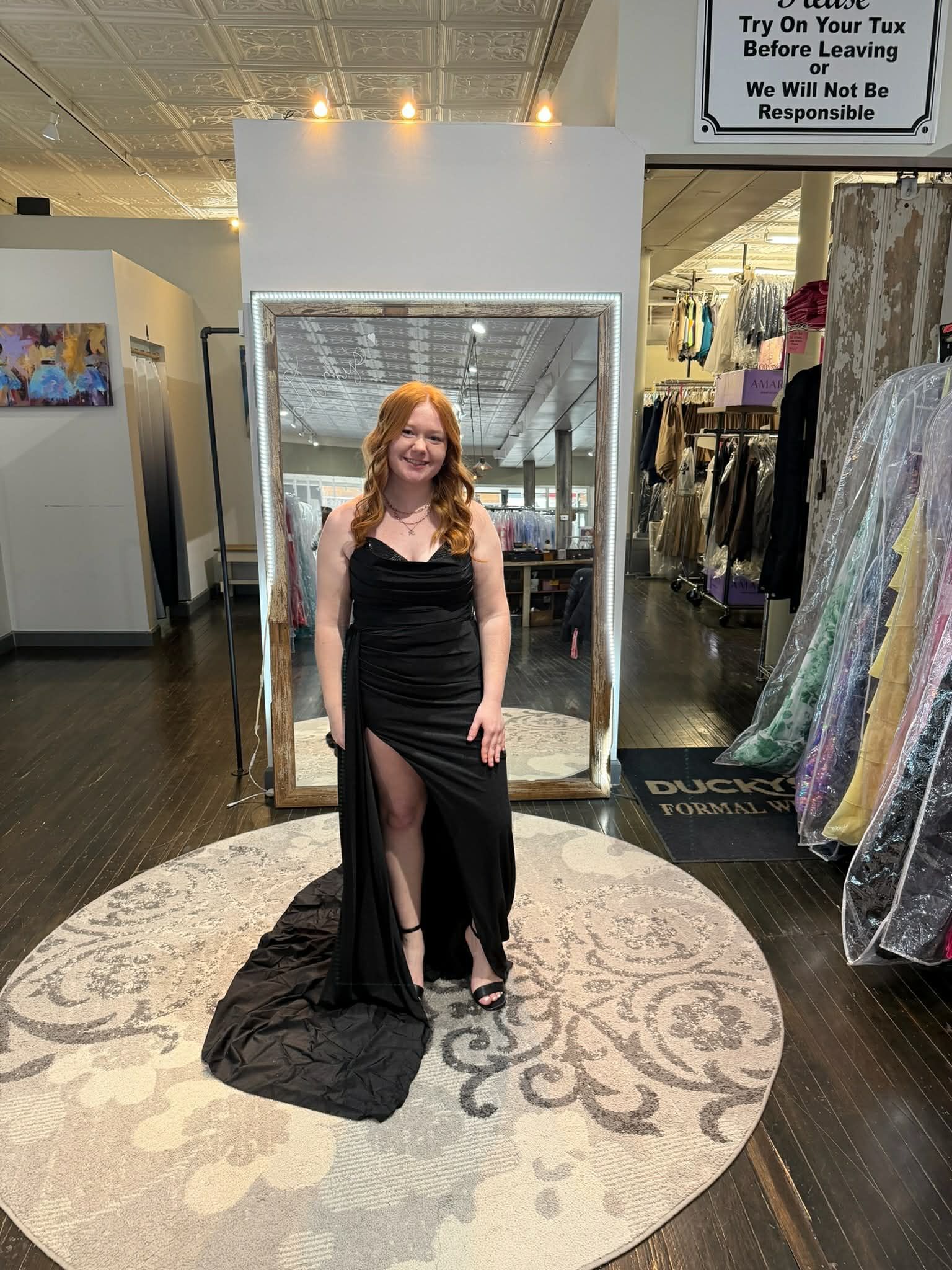 A person in a black floor-length formal dress with a high side slit stands on a round rug in a boutique store.