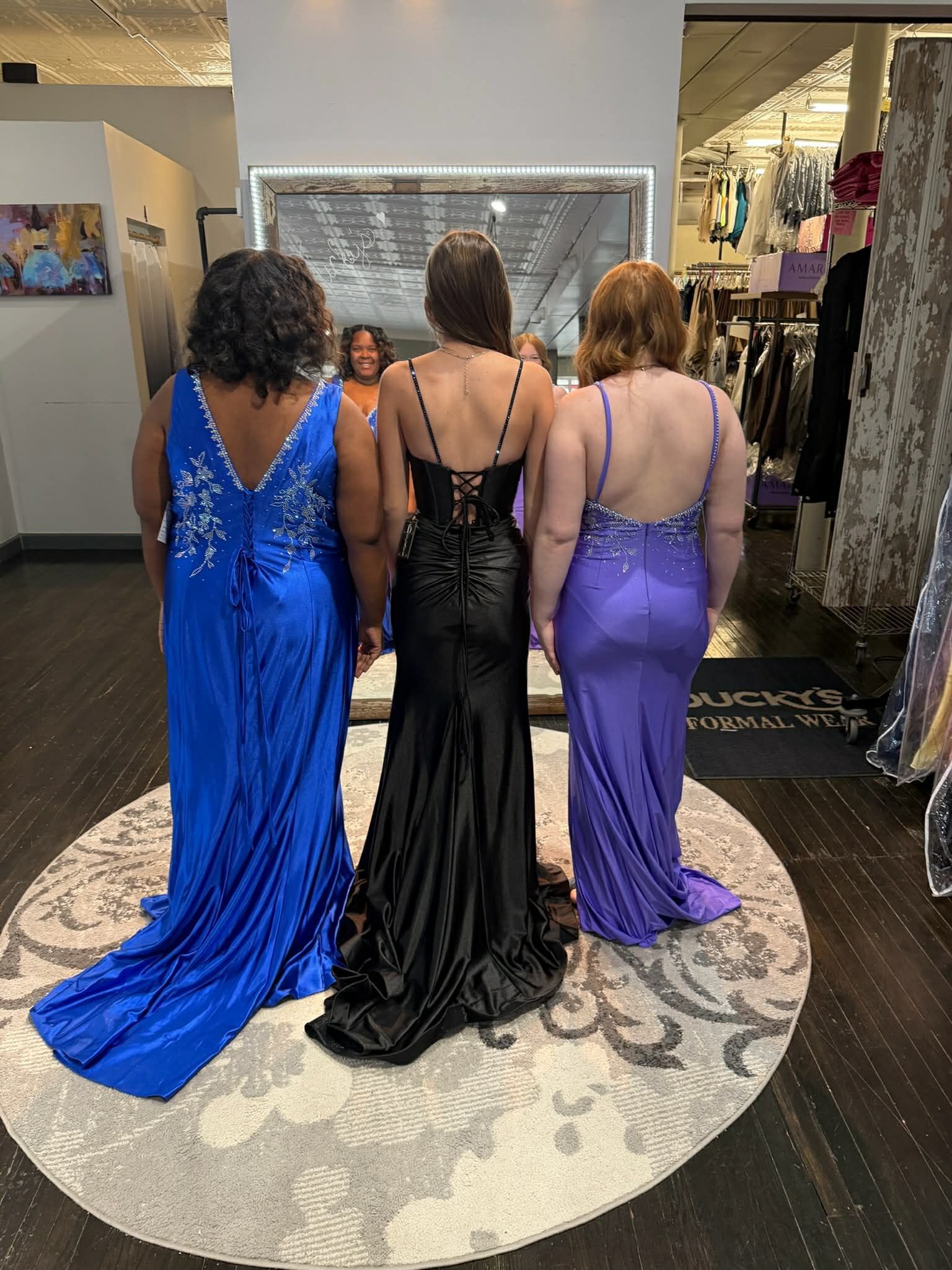 Three people stand in a clothing store wearing floor-length evening gowns in blue, black, and purple.