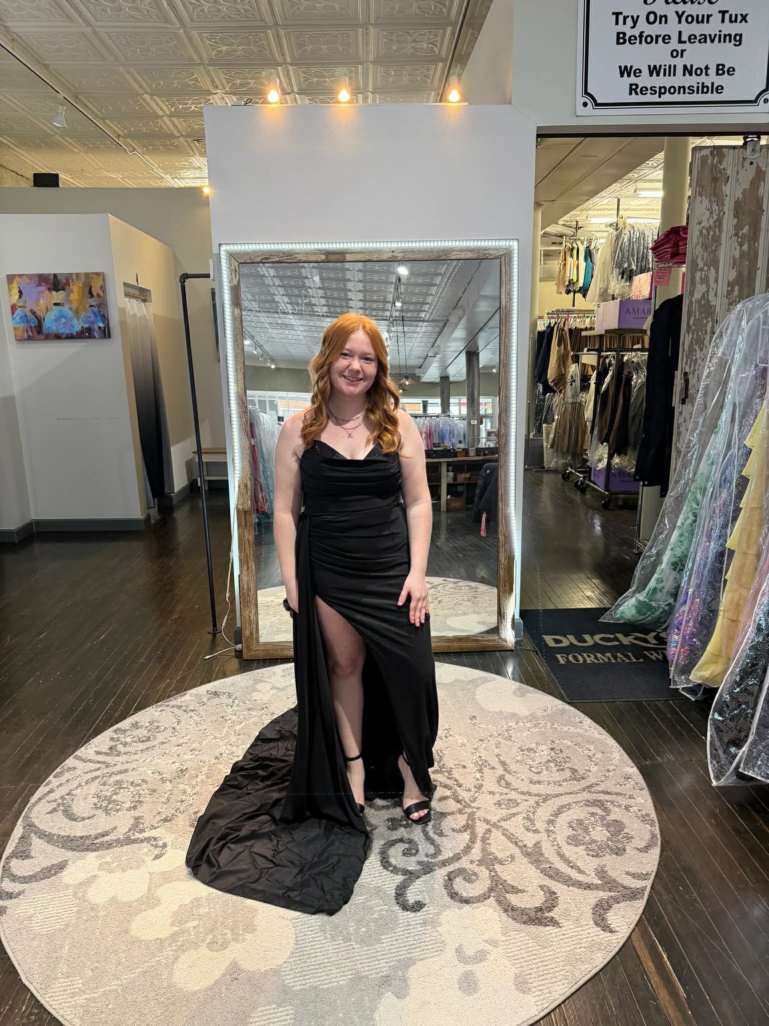 A smiling person stands on a circular rug in a formal wear shop, wearing a floor-length black dress with a side slit.