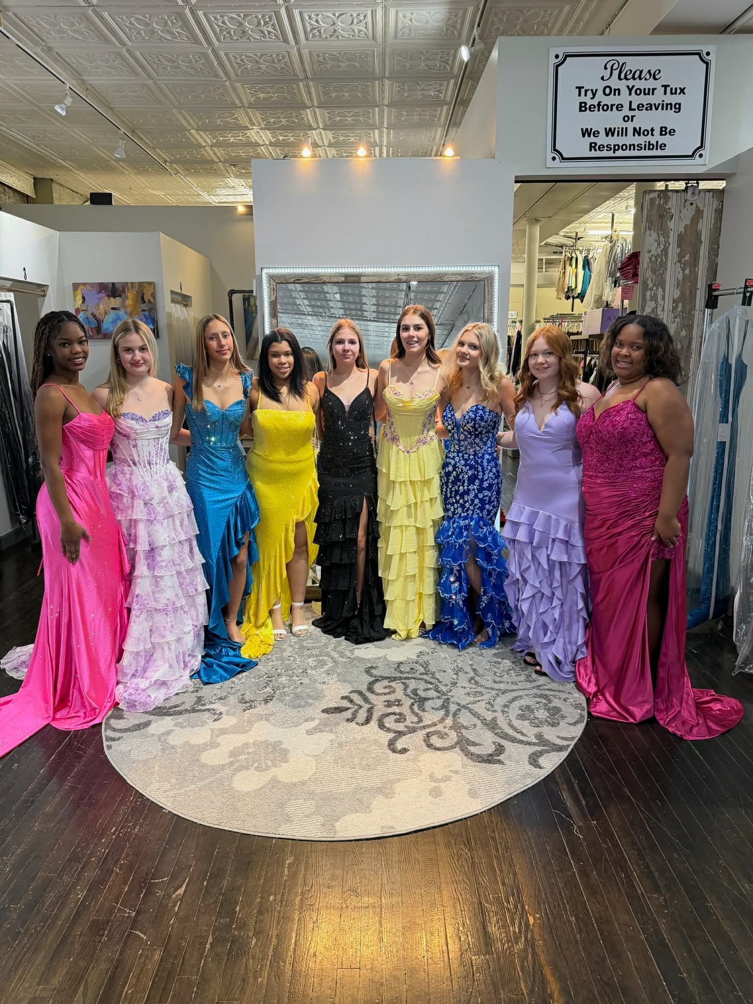 Eight people stand in a line in a store, wearing long, colorful, formal dresses of various styles.