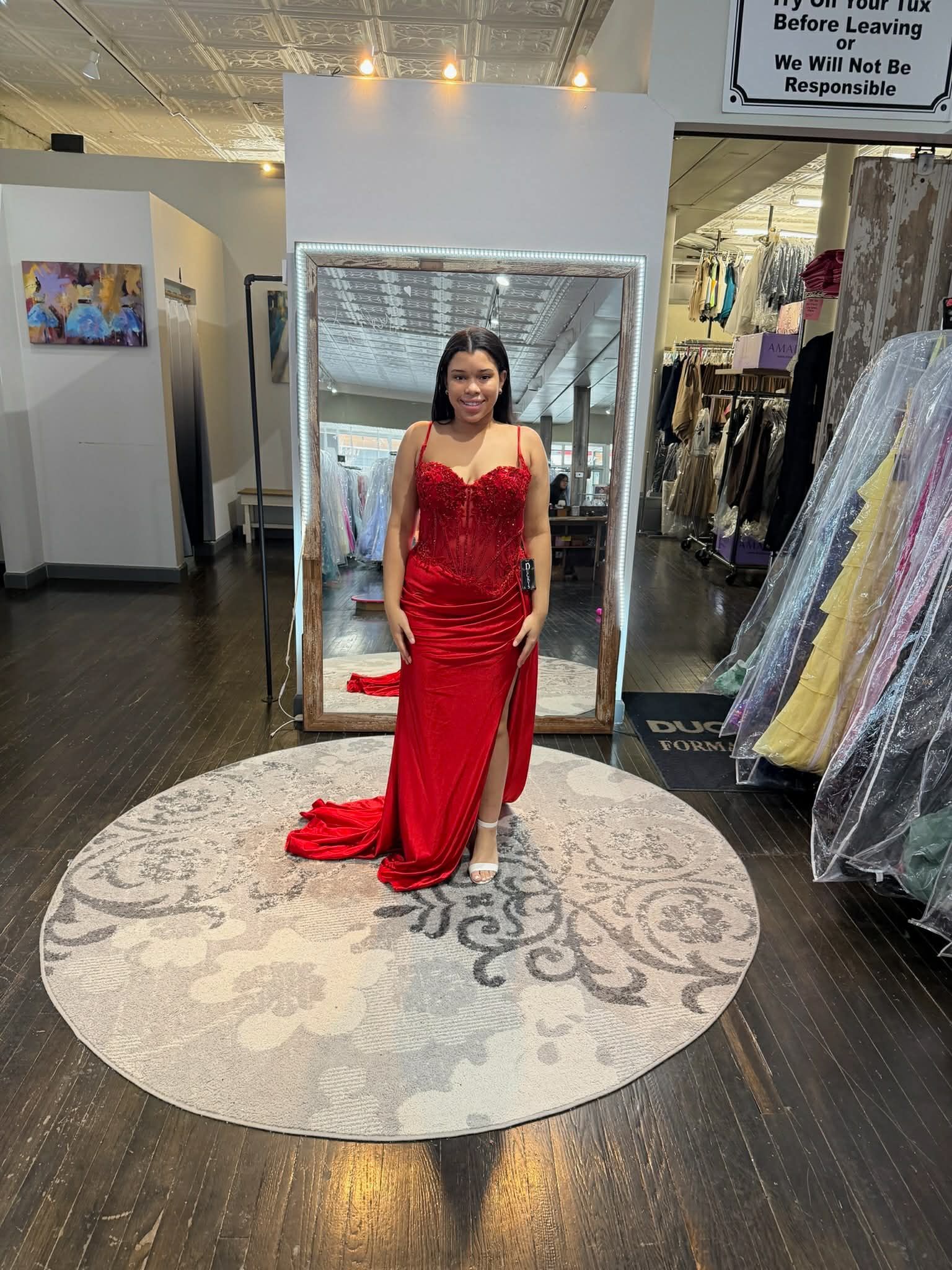 A person wears a red, floor-length sequined evening gown with a high slit, standing on a round rug in a clothing boutique.