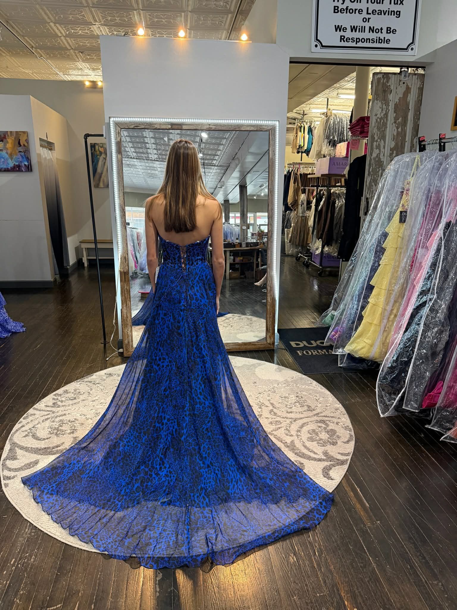 A person stands in a dress shop wearing a floor-length, deep blue strapless gown with an intricate lace train.