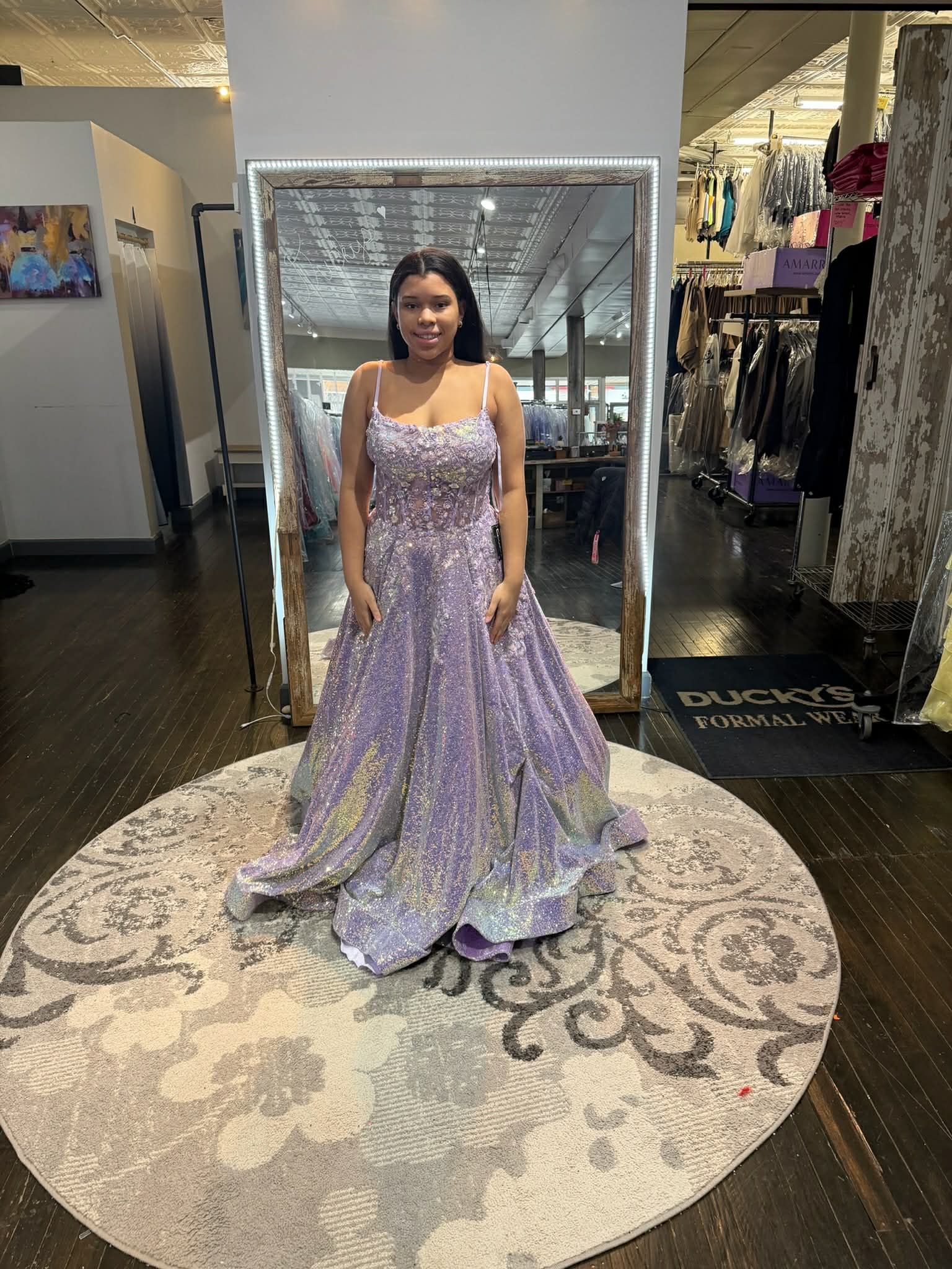 A person stands in a boutique wearing a floor-length, shimmering, lavender sequined gown with spaghetti straps.