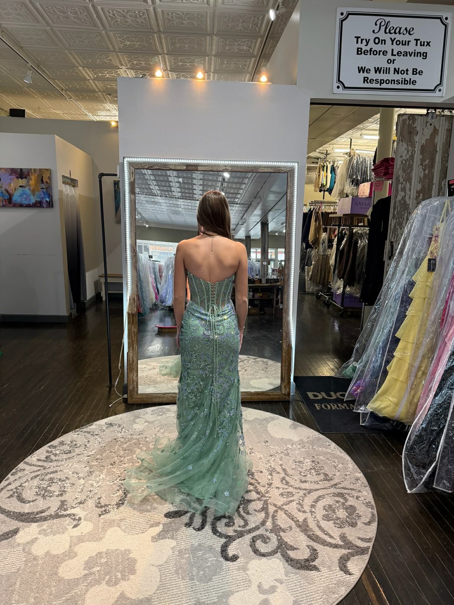 A person stands in a dress shop before a mirror, wearing a backless, light green, sequined evening gown with a small train.