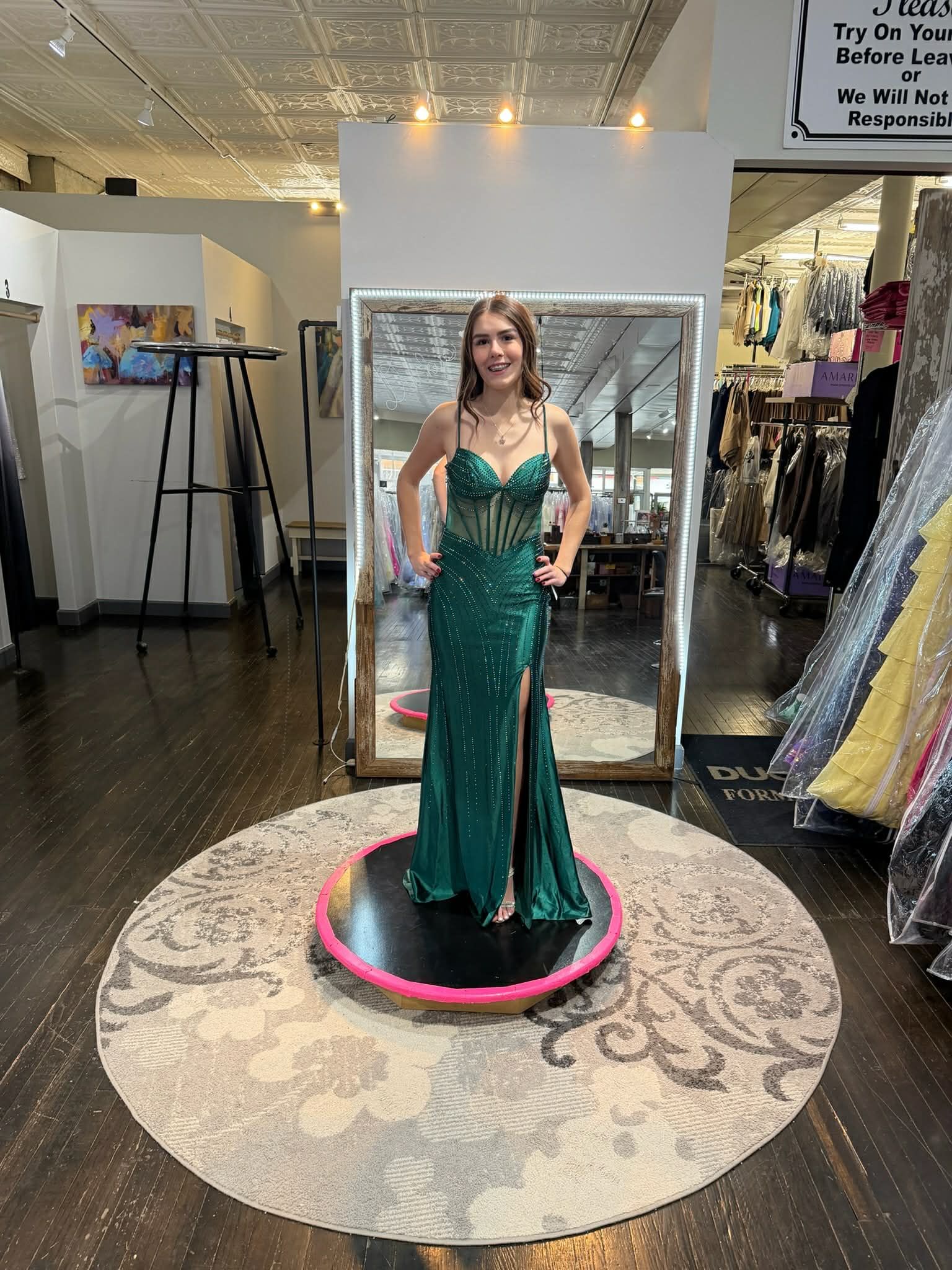 A person in a floor-length, emerald-green gown with a high slit, standing on a circular platform in a clothing store.