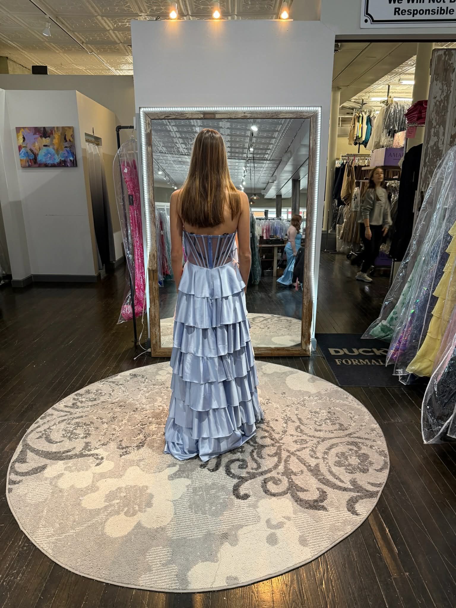 A person stands in a dress shop, facing a mirror, wearing a floor-length, light blue, tiered ruffled strapless gown.
