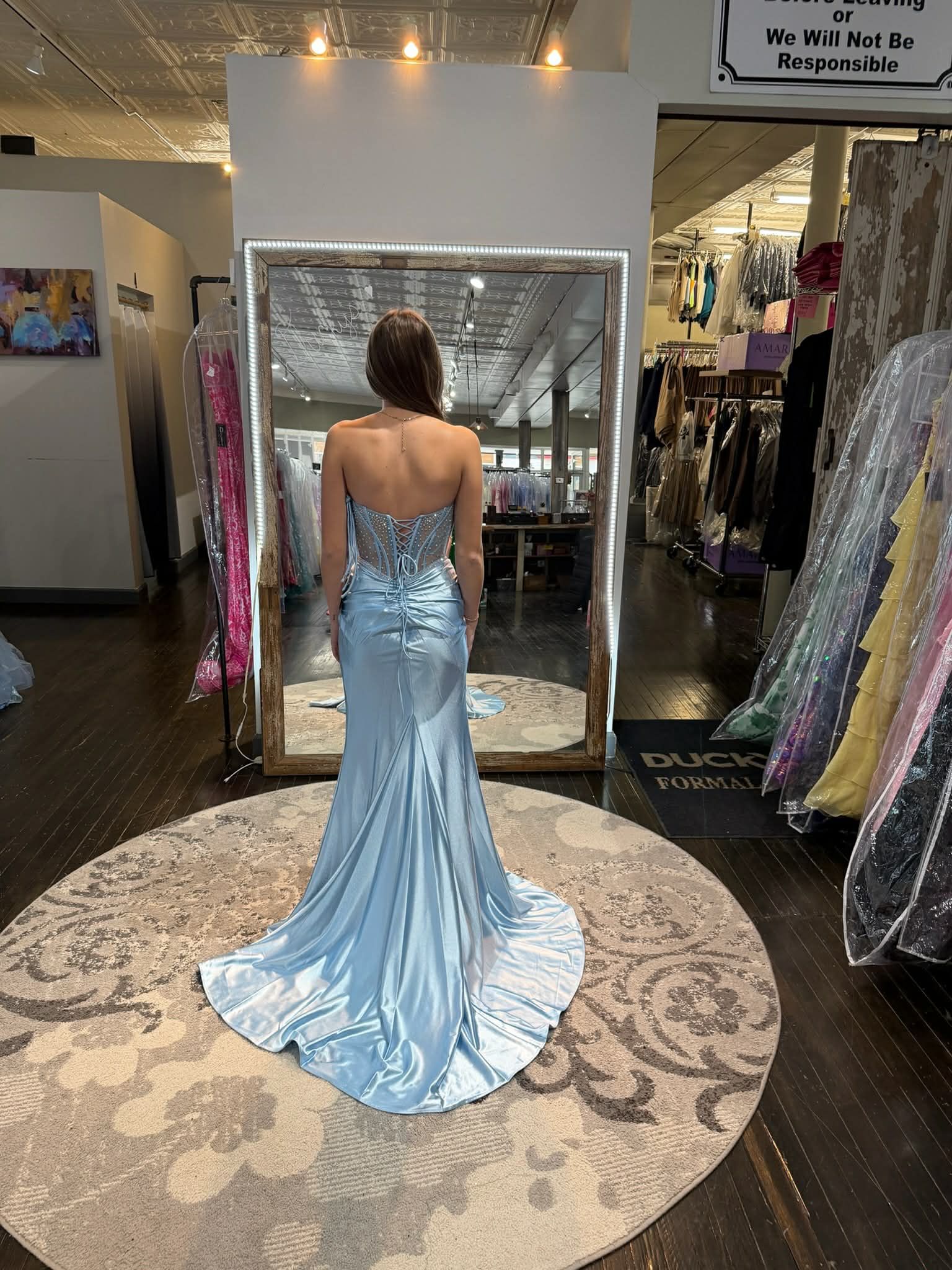 A person stands in a dress shop, facing a mirror, wearing a light blue, strapless gown with a multi-tiered ruffled skirt.