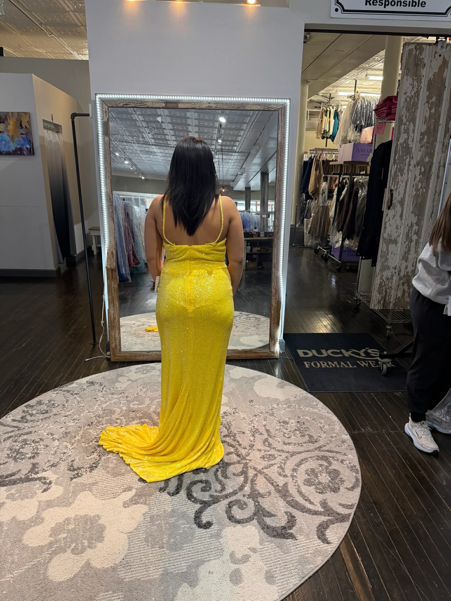 A person stands in a dress shop, facing a mirror and wearing a bright yellow, floor-length sequined evening gown.