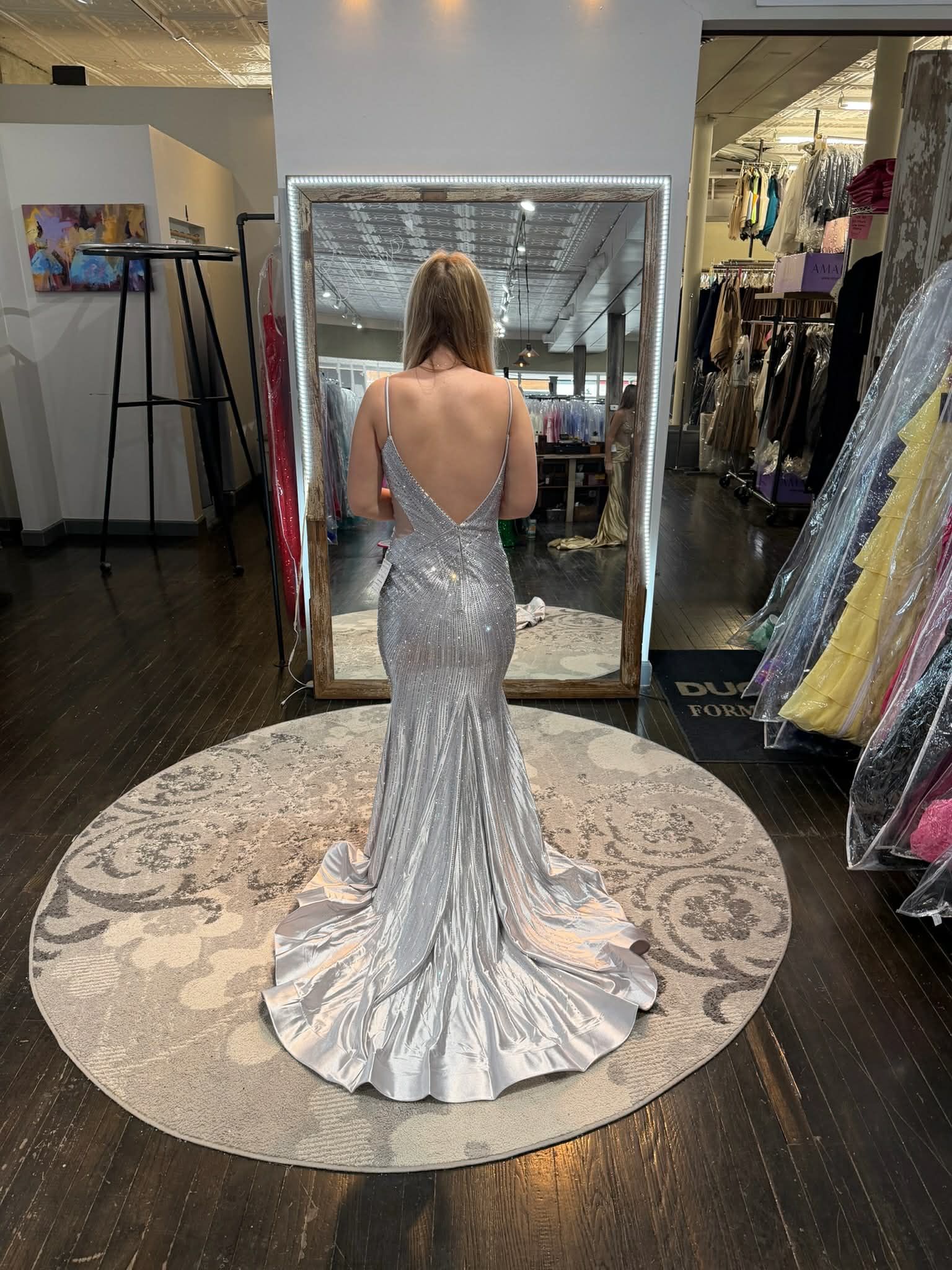 A person stands on a circular rug in a boutique, wearing a backless, silver sequined gown with a flared train.