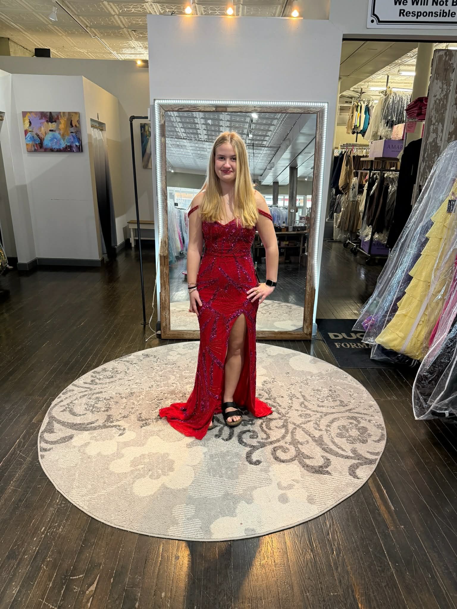 A person wearing a long, red sequined dress with a thigh-high slit, standing on a round patterned rug in a clothing store.