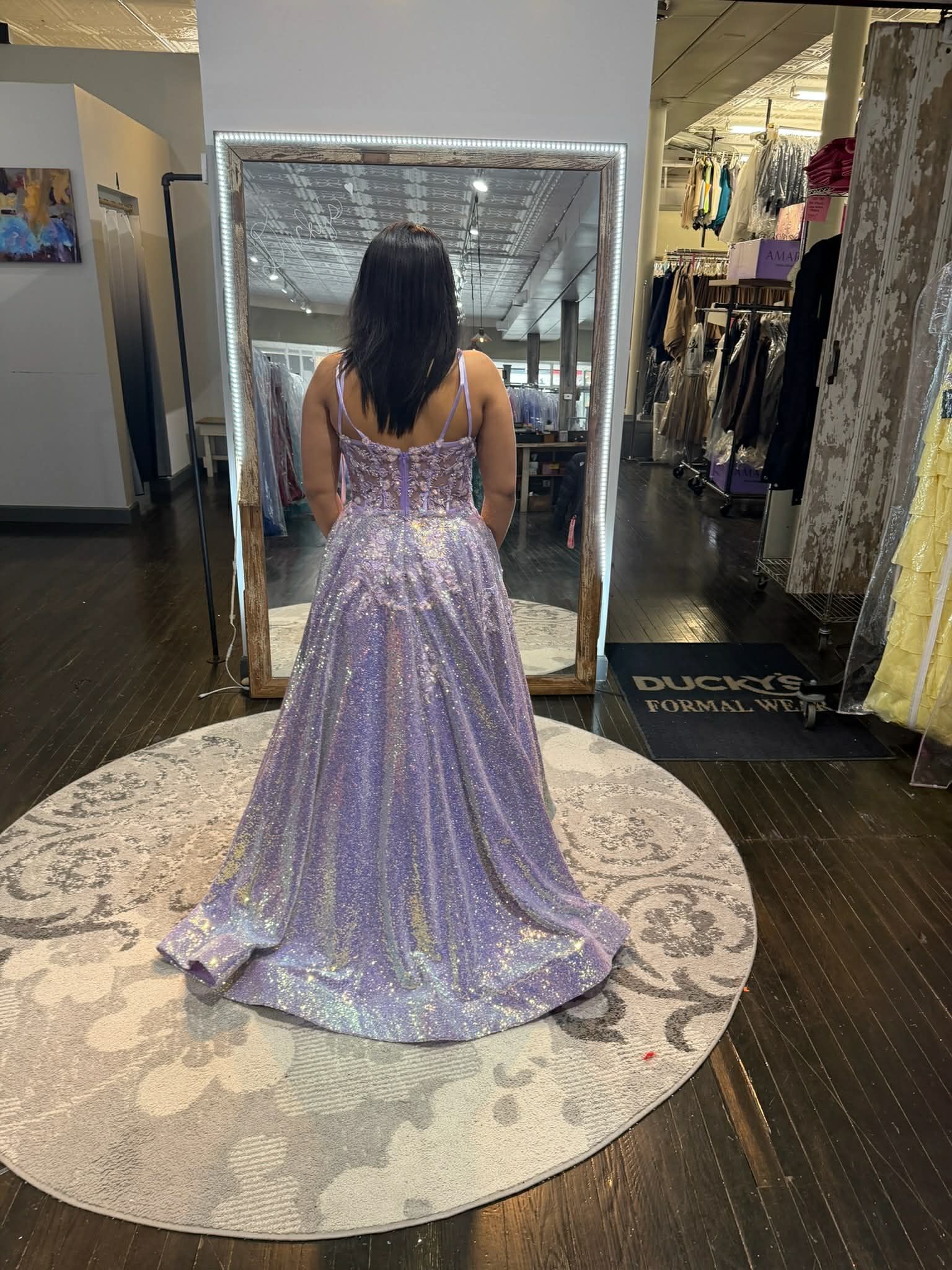 A person standing on a round rug in a store, wearing a floor-length lavender sequined gown viewed from the back.