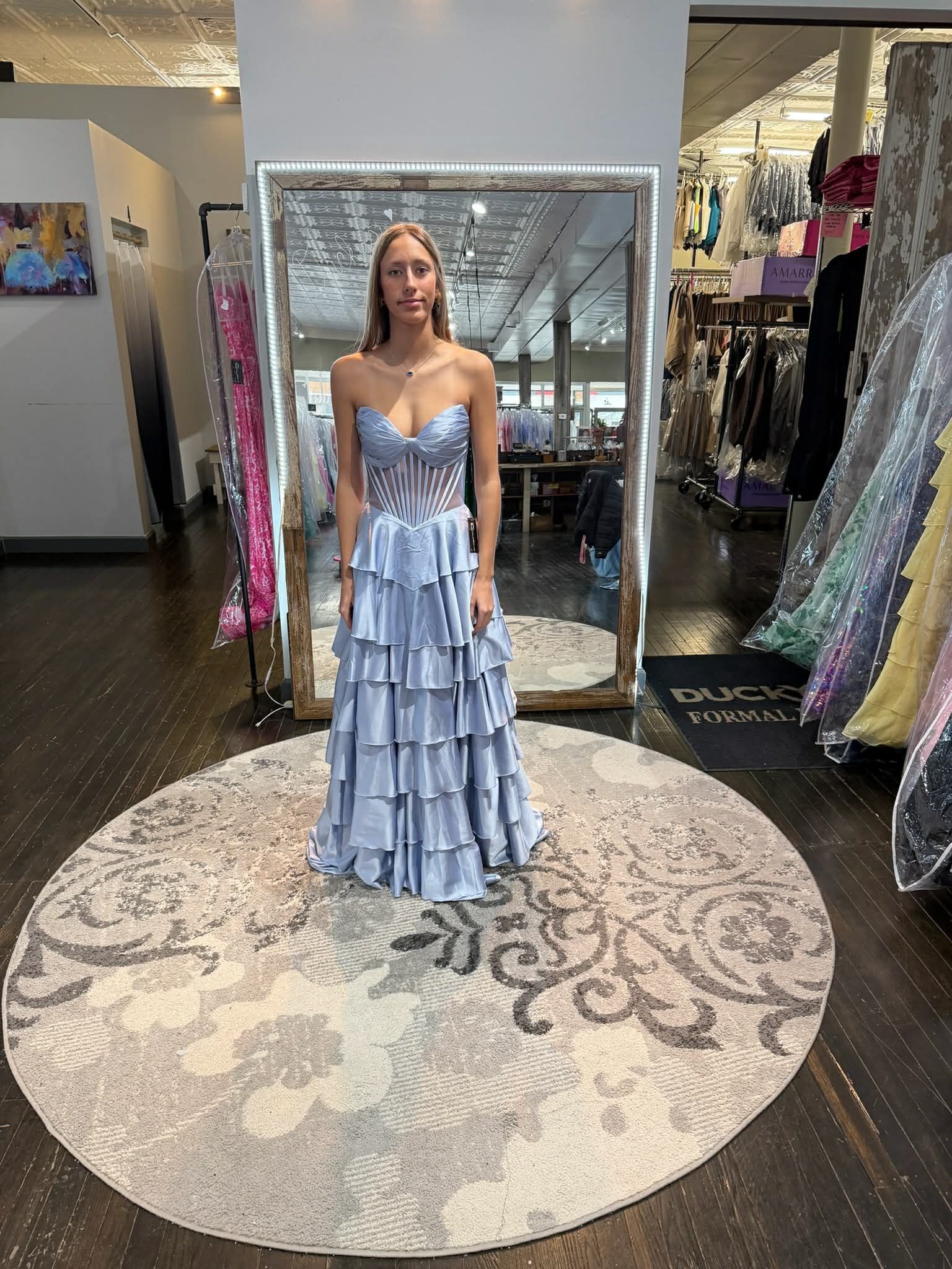 A person stands in front of a mirror wearing a light blue, strapless, tiered ruffle gown in a clothing store.