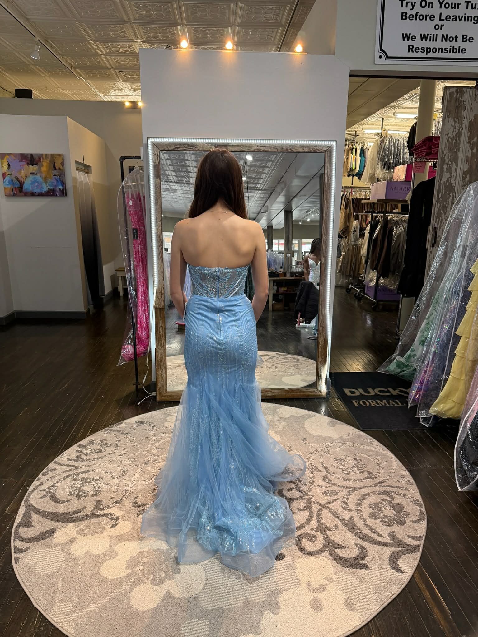 A person stands in a dress shop wearing a floor-length, strapless, light blue sequined gown with a mermaid-style skirt.