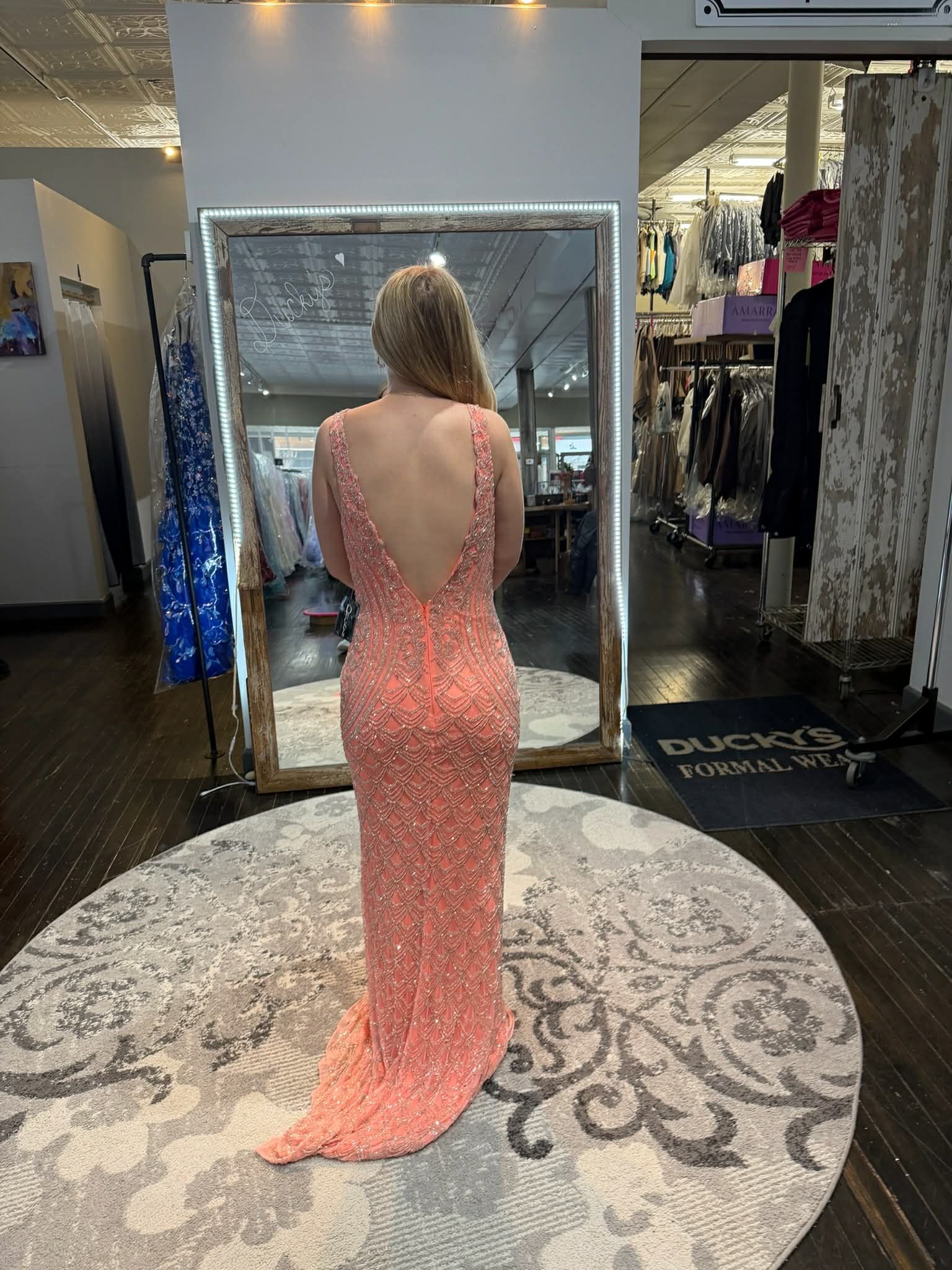 A person stands in a dress shop, seen from the back, wearing a floor-length, backless coral sequined gown.