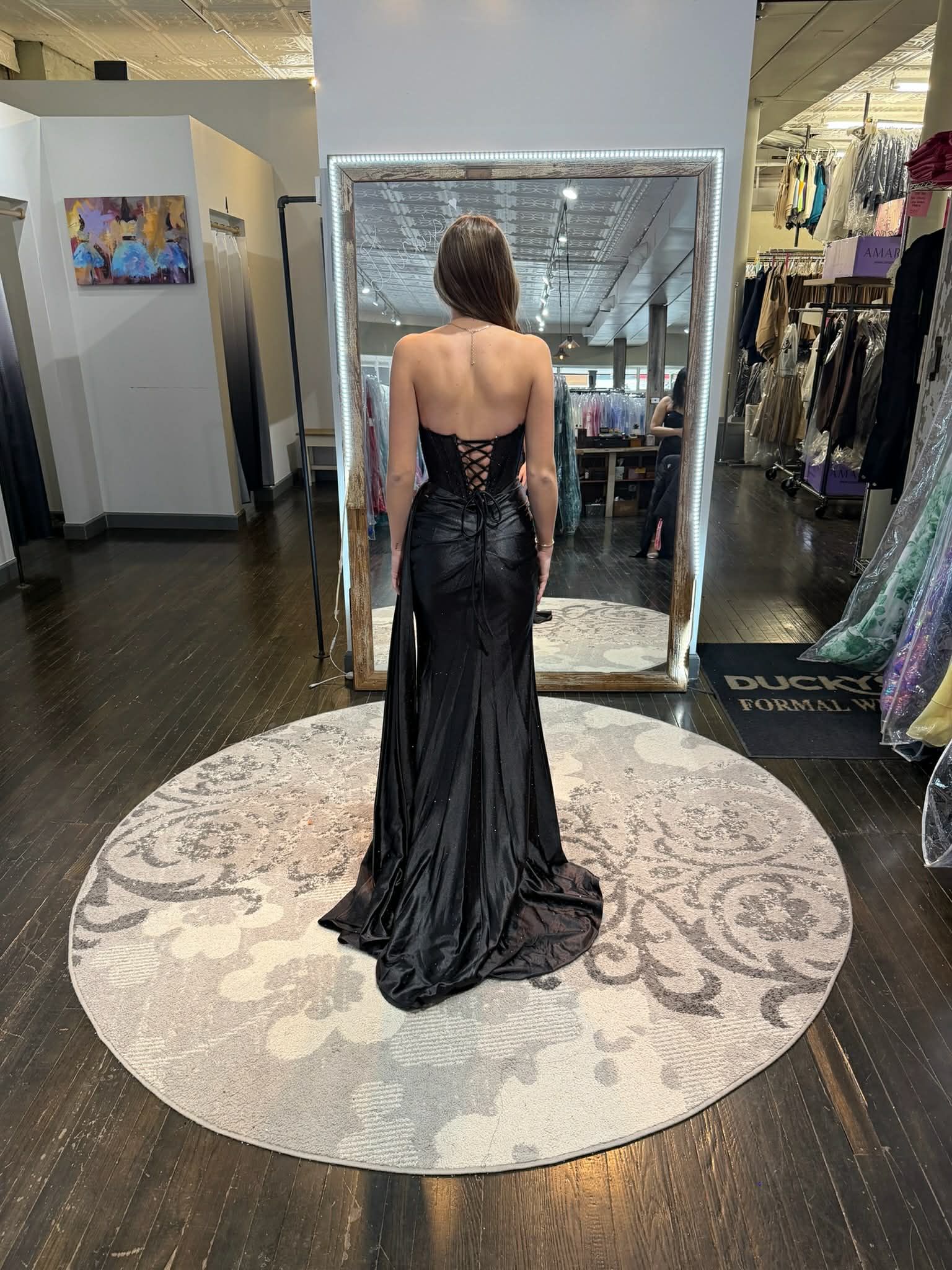 A person wearing a black, strapless, lace-up corset gown with a train, standing on a circular rug in a formal wear store.