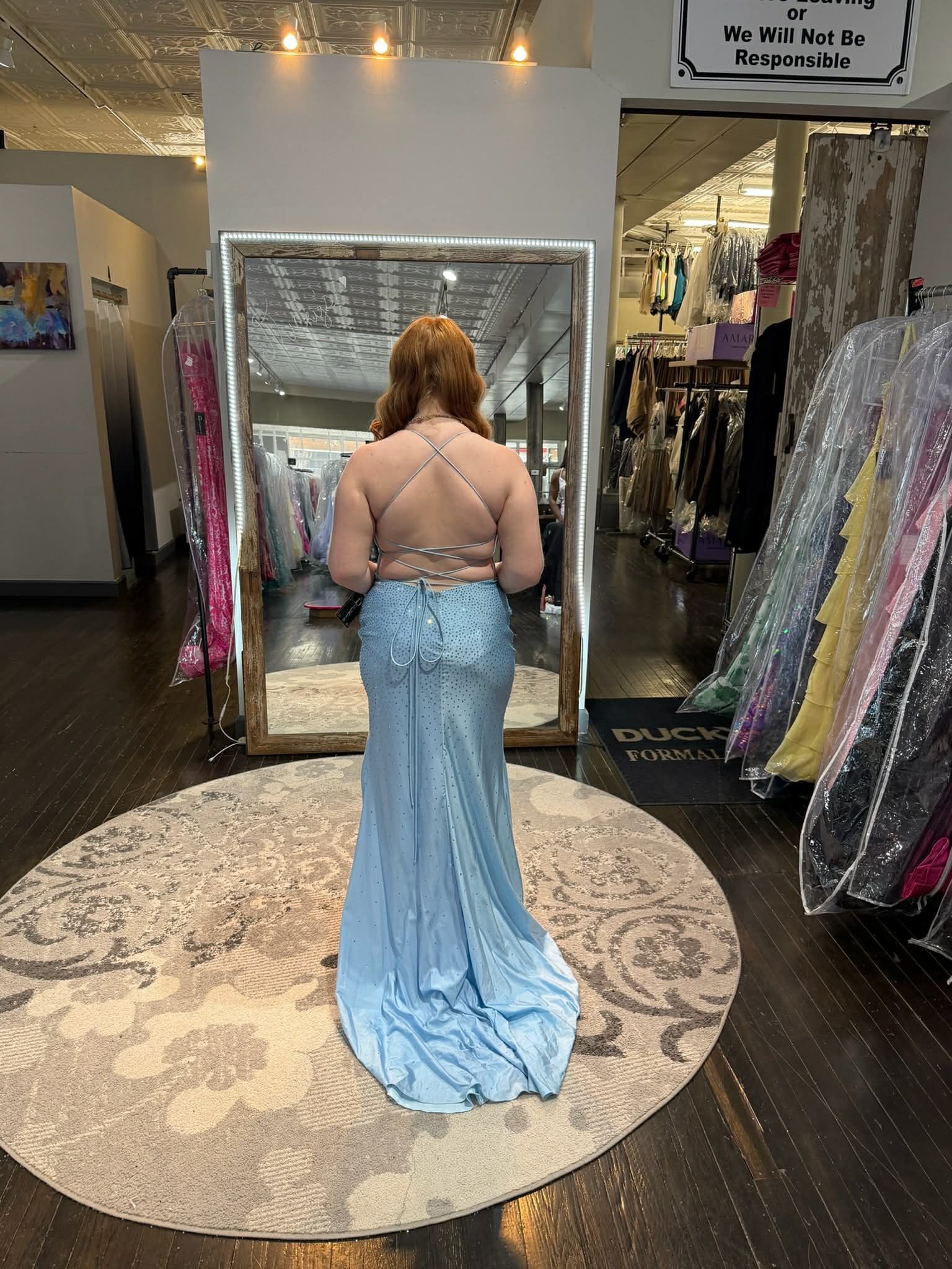 A person stands in a dress shop wearing a backless, light blue floor-length gown with a train, facing a large mirror.