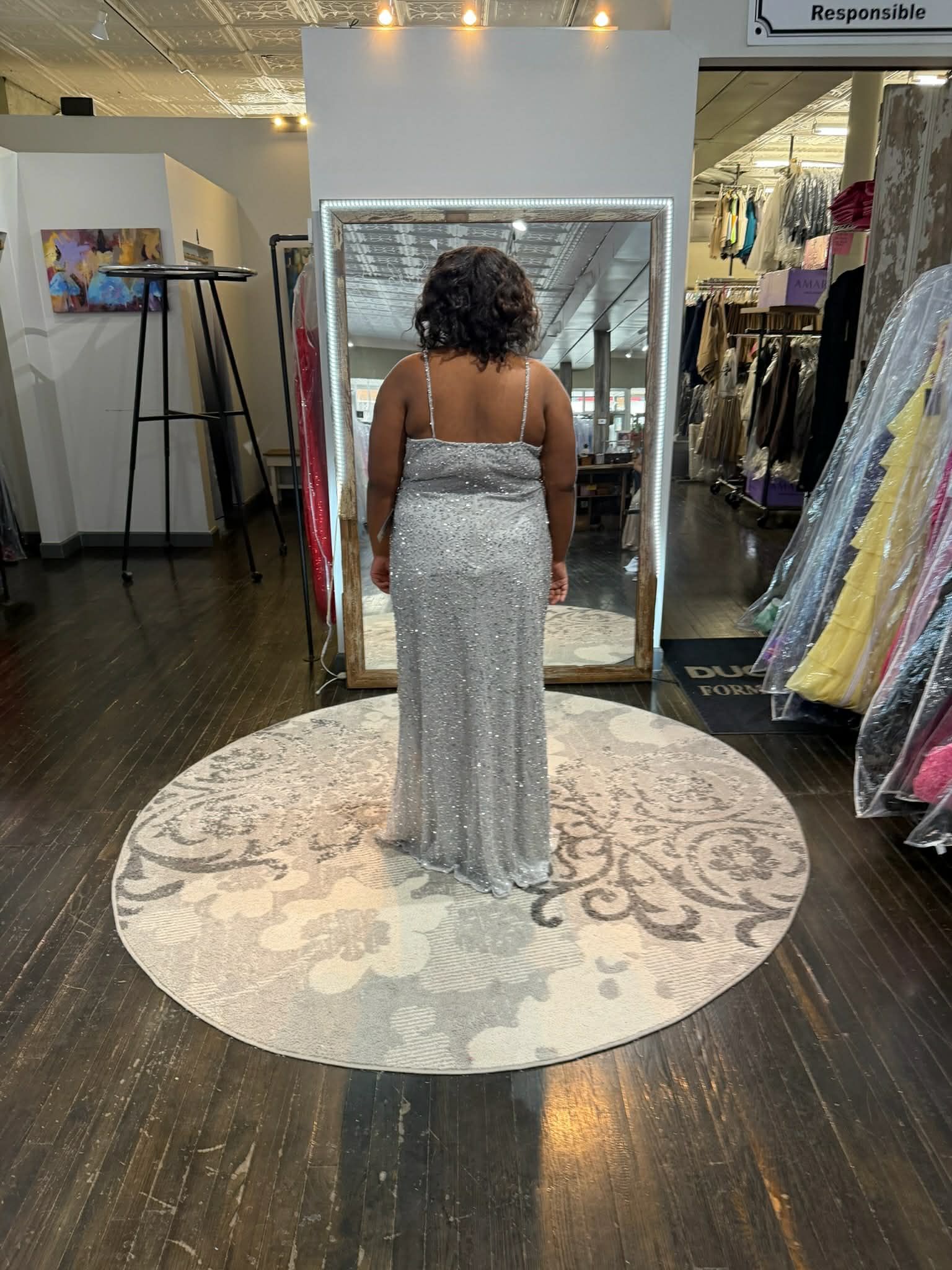 A person stands on a circular rug in a boutique, wearing a floor-length, silver sequined evening gown viewed from behind.