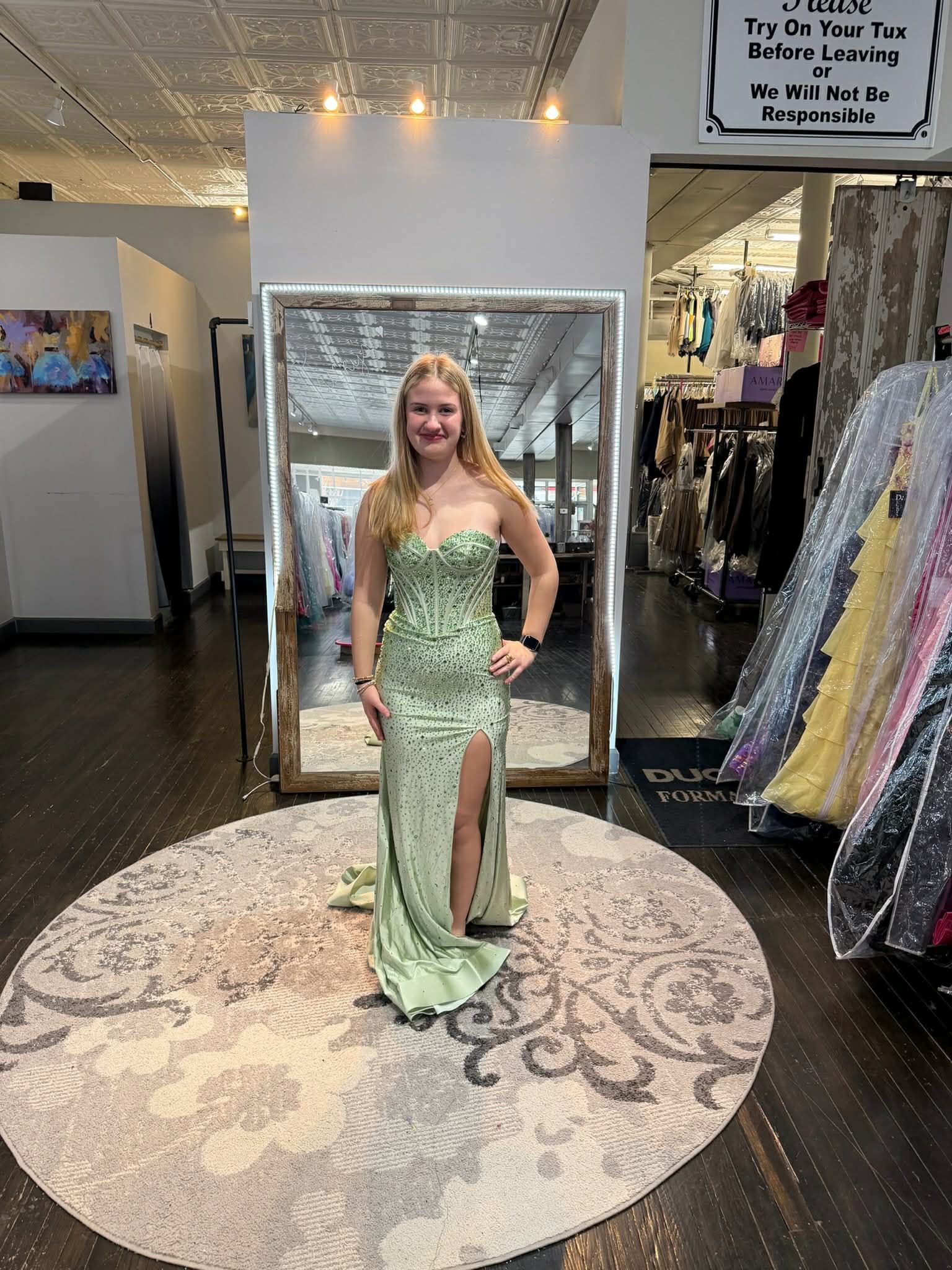 A person smiles while standing on a circular rug in a clothing store, wearing a floor-length, pale green floral gown.