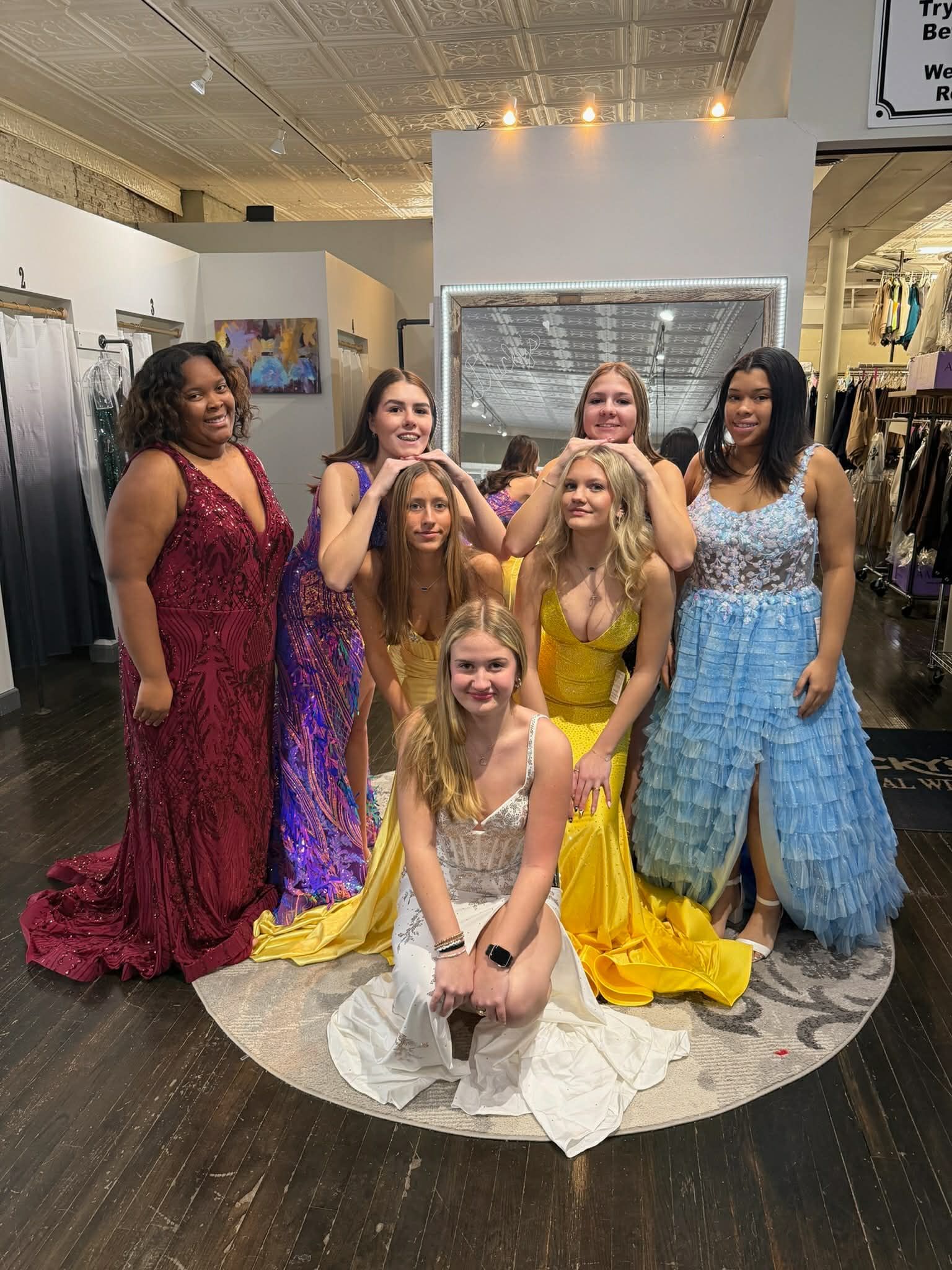 Six people pose in a boutique, wearing formal gowns in red, purple, white, and yellow, with one person kneeling in front.