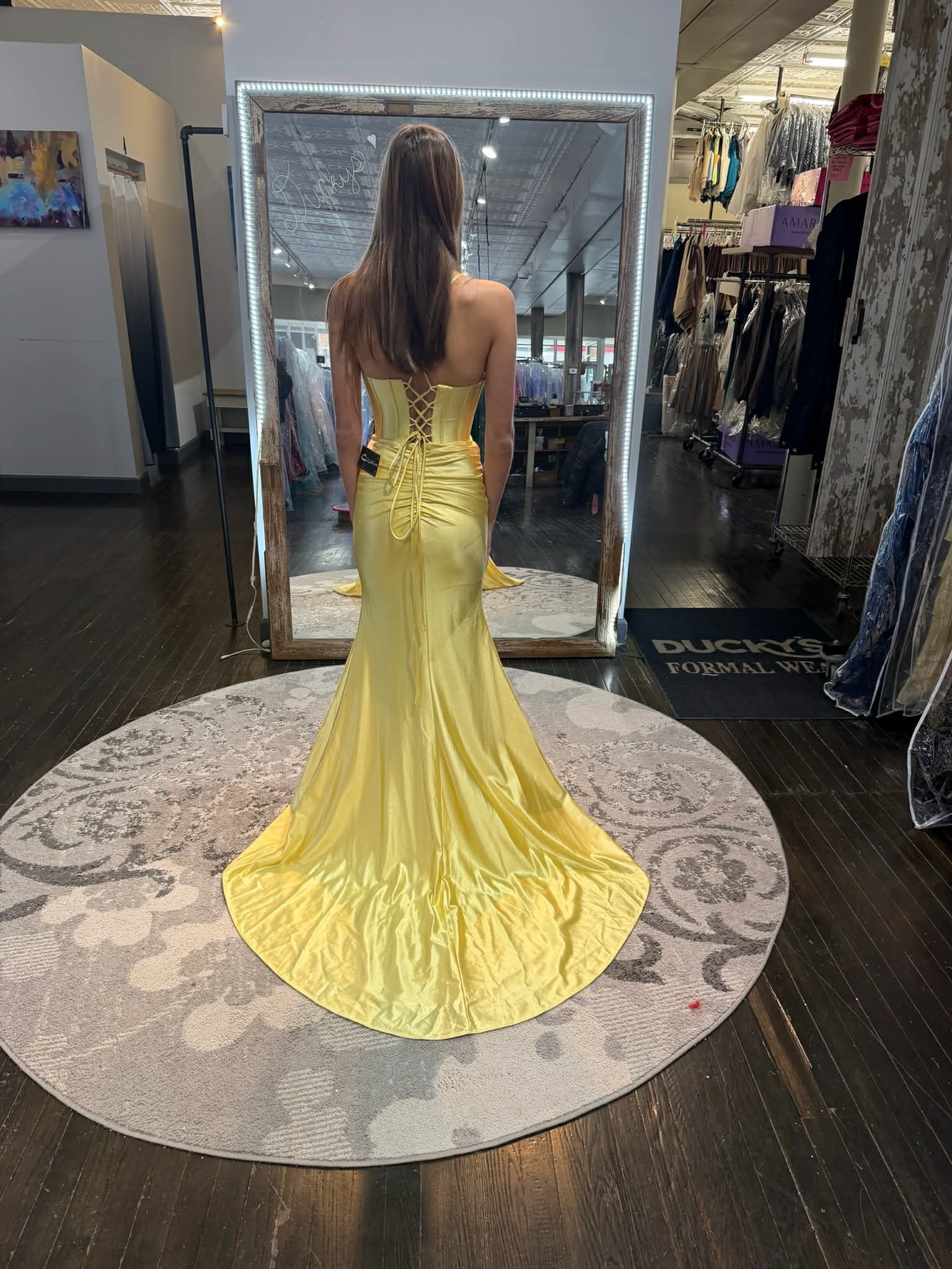 A person stands on a round rug in a clothing store, facing a mirror while wearing a floor-length, yellow, mermaid-cut gown.