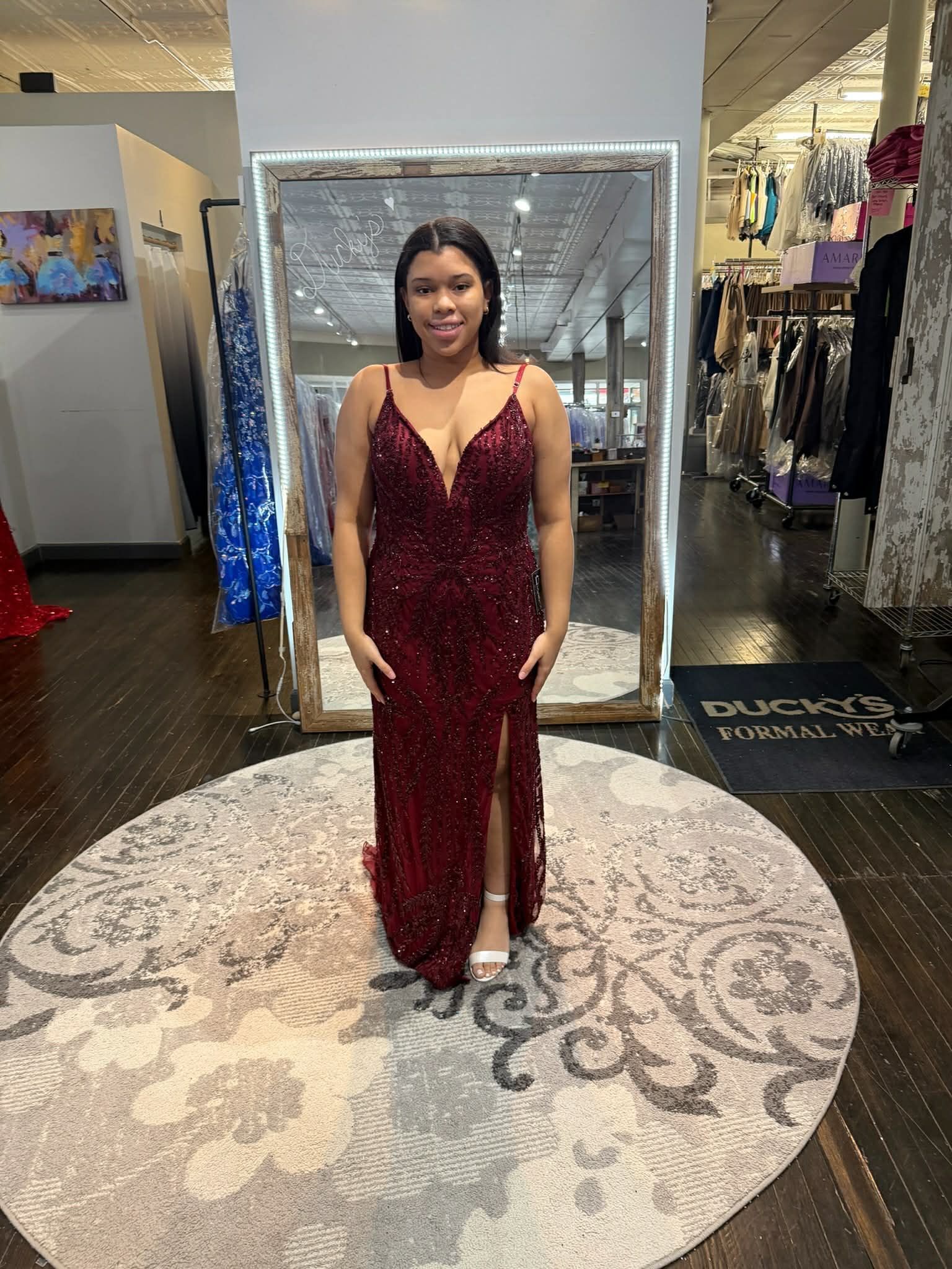 A person in a floor-length, deep red sequined gown with a leg slit stands on a circular rug in a clothing store.