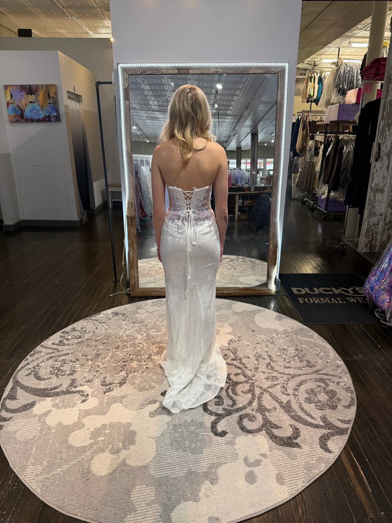 A person stands in a clothing store, seen from behind while wearing a sequined, lace-up back evening gown in a mirror.