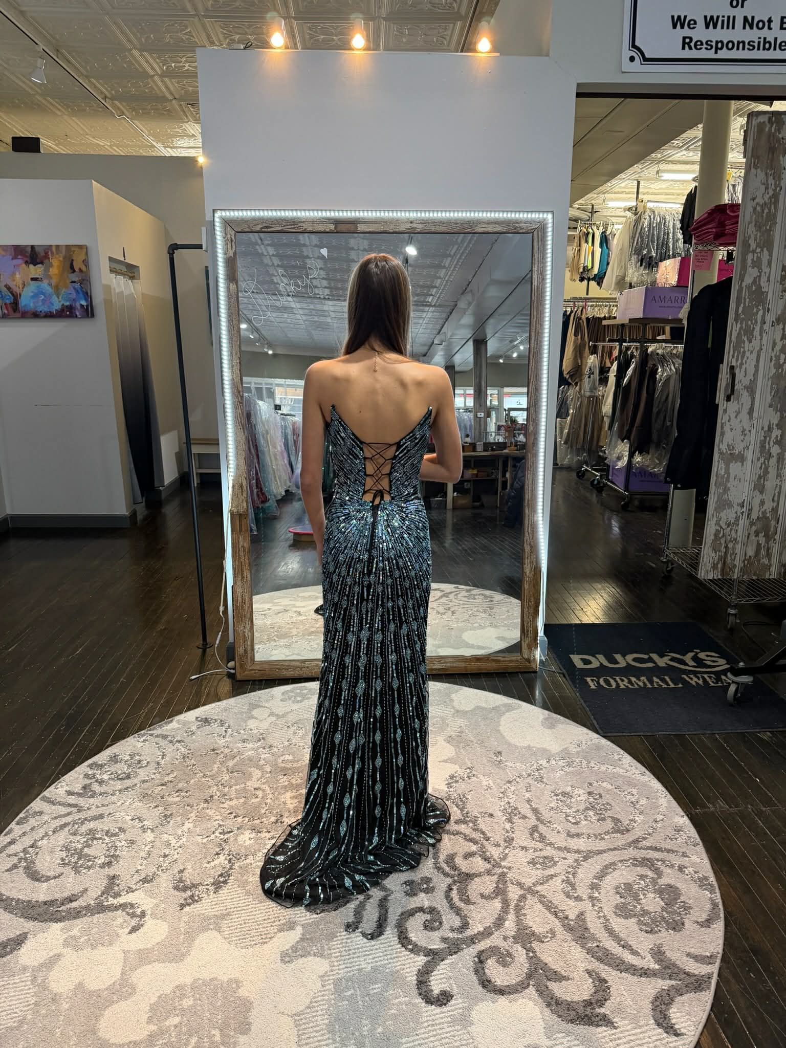 A person standing on a small platform in a clothing store, modeling a long, shimmering, dark green sequined gown.