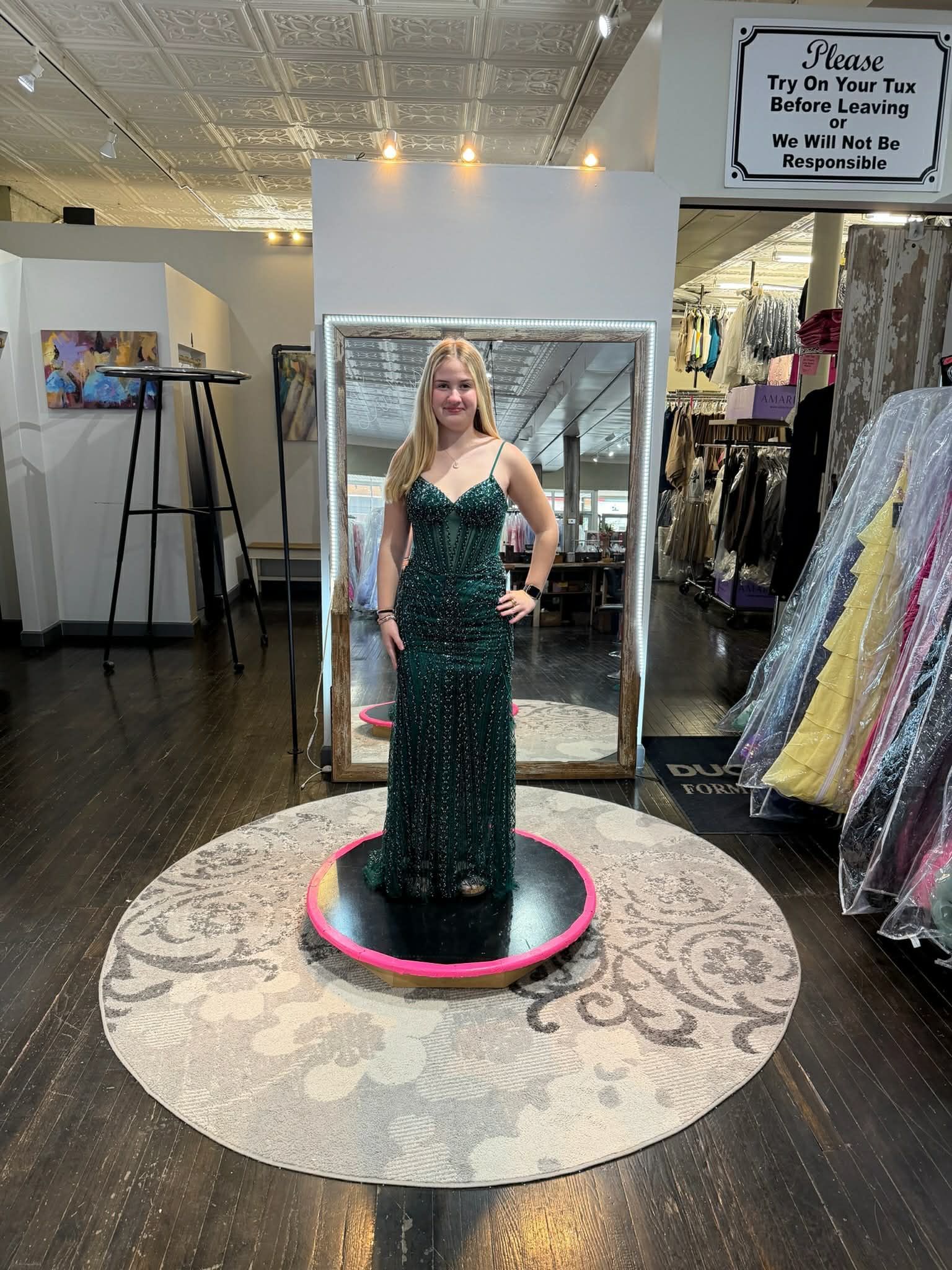 A person stands in a clothing store, facing a large mirror while wearing a floor-length, backless, coral sequin gown.