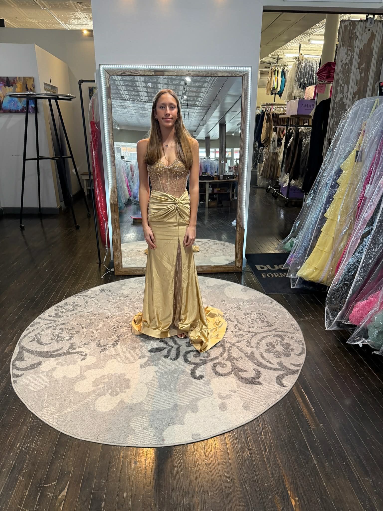 A person wearing a shimmering blue, ruffled high-low formal gown stands on a round patterned rug in a clothing boutique.