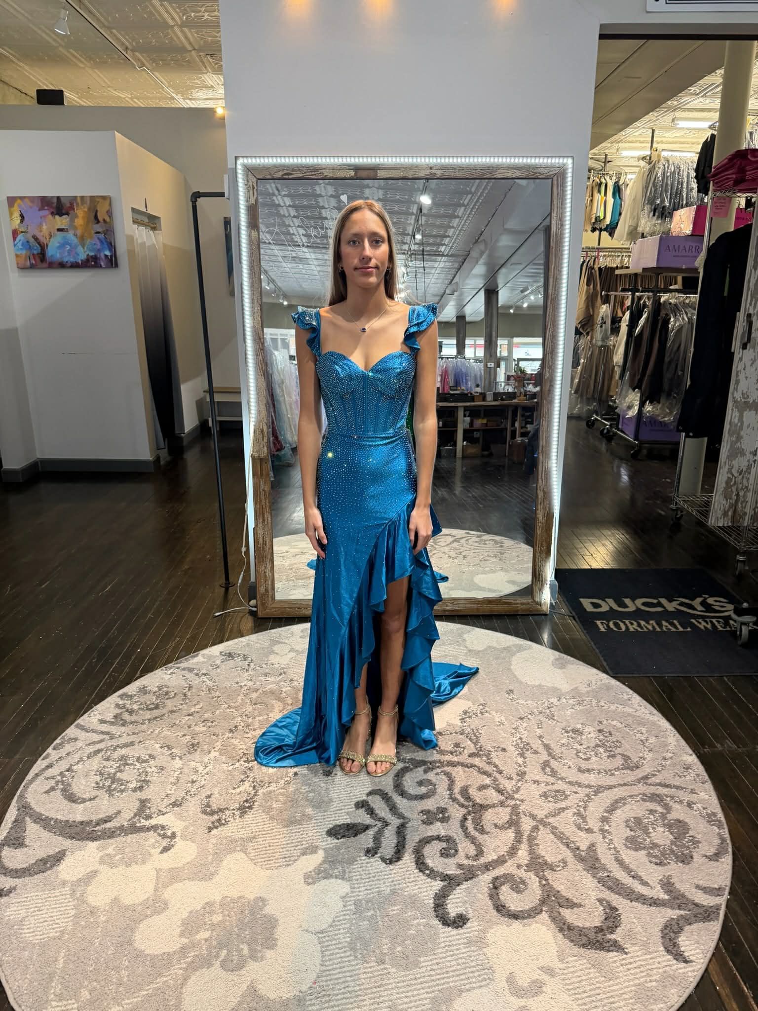 A person wearing a long, sparkling red dress with an open back, standing on a circular rug in front of a floor mirror.