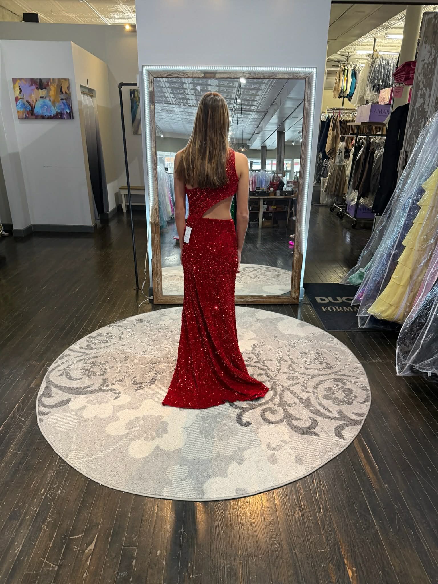 A person stands on a round rug in a clothing boutique, wearing a long, blue, strapless, sequined gown with a leg slit.