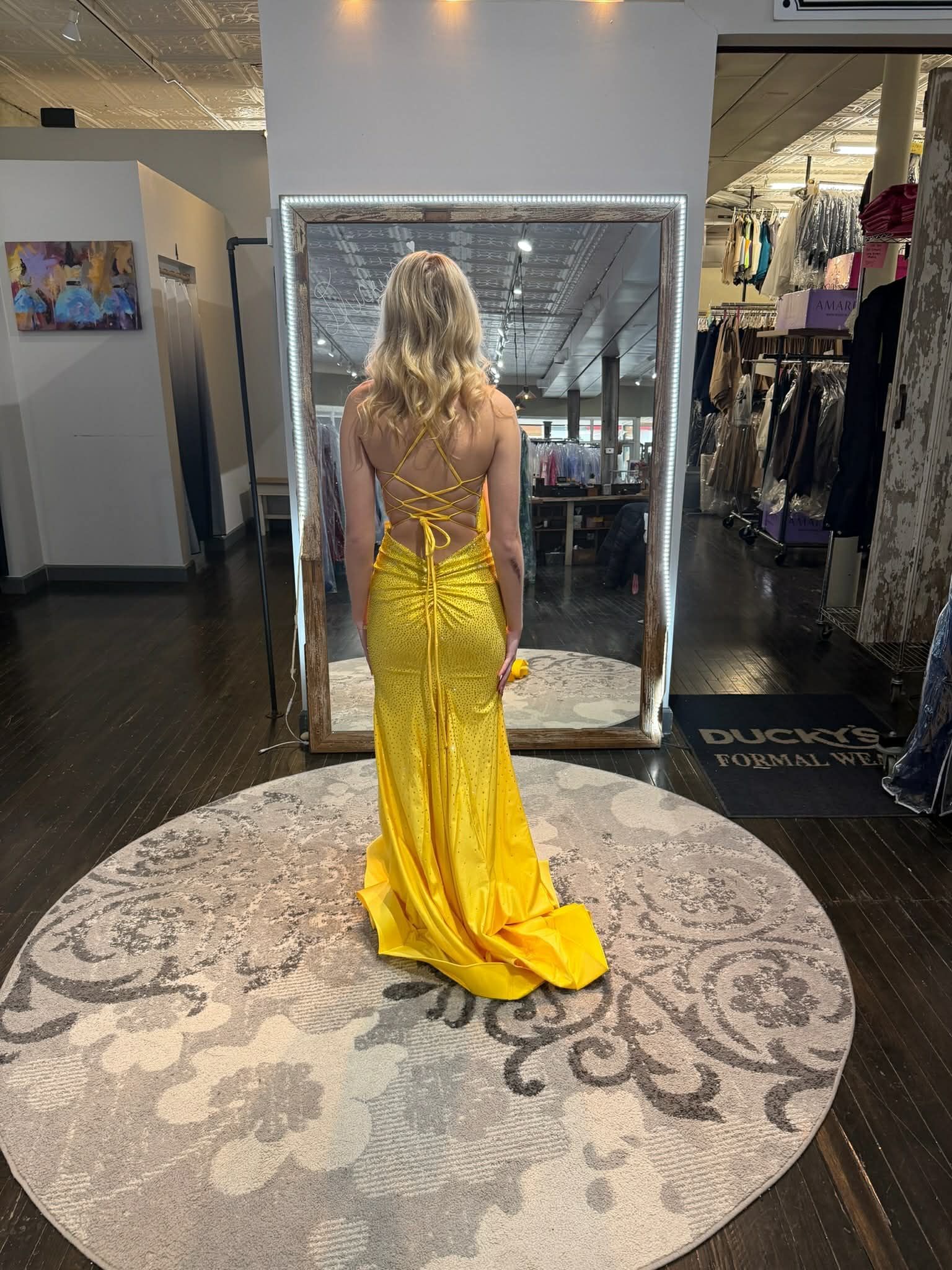 A person stands in a clothing store, facing a mirror while wearing a white floor-length gown with pink floral patterns.