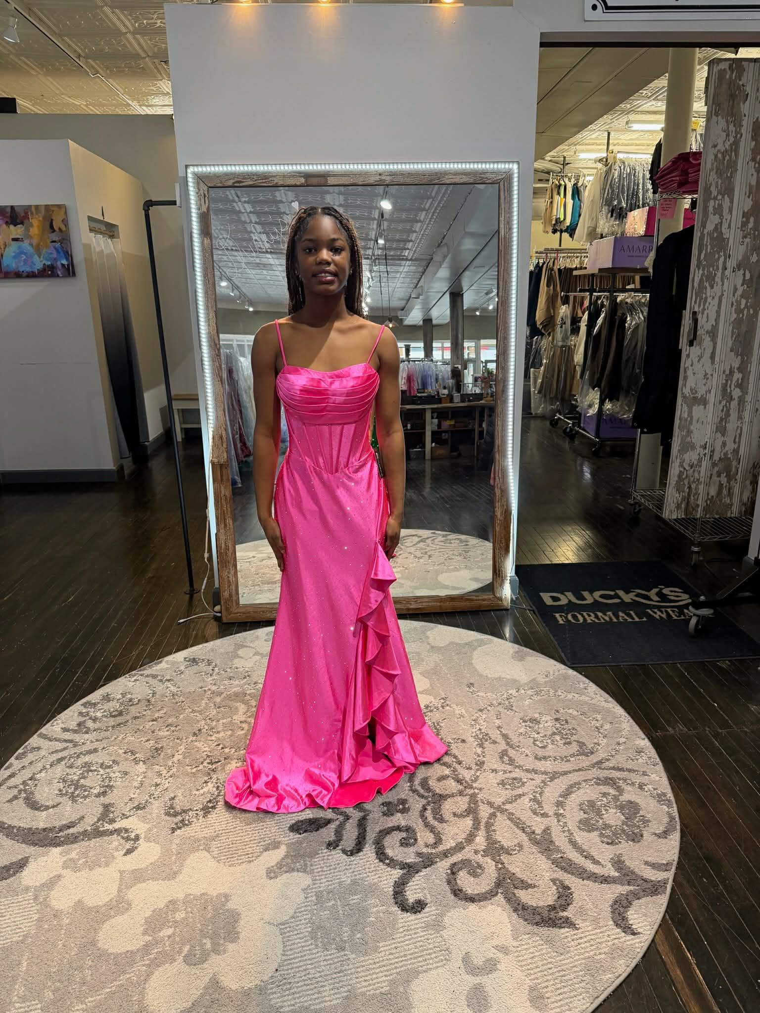 A person stands on a circular rug in a boutique, wearing a bright pink, floor-length gown with a ruffled side slit.