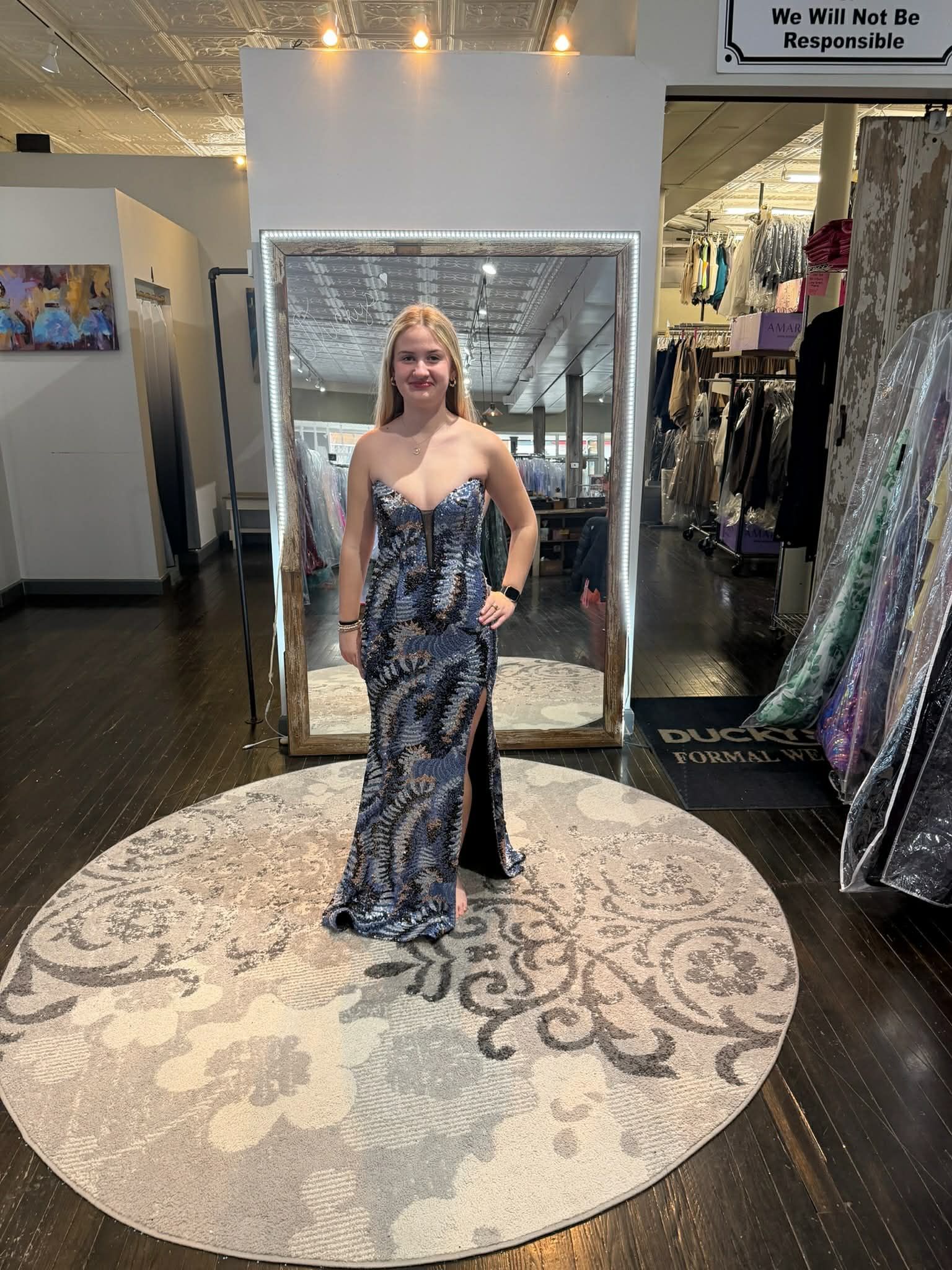 A person smiles while standing on a rug in a store, wearing a floor-length, strapless blue and black patterned gown.