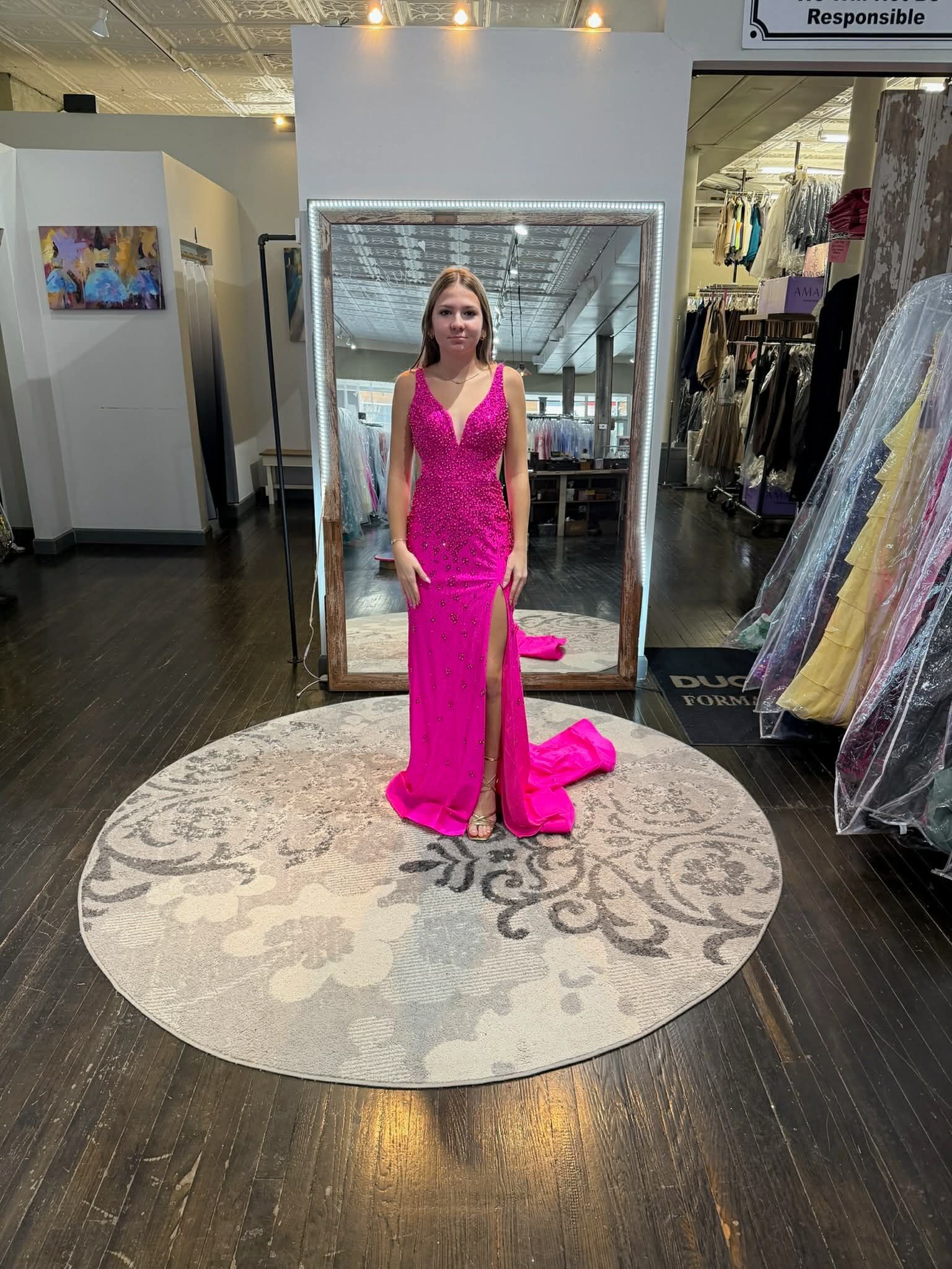 A person standing on a round rug in a clothing boutique wearing a floor-length, bright pink gown with a corset back.